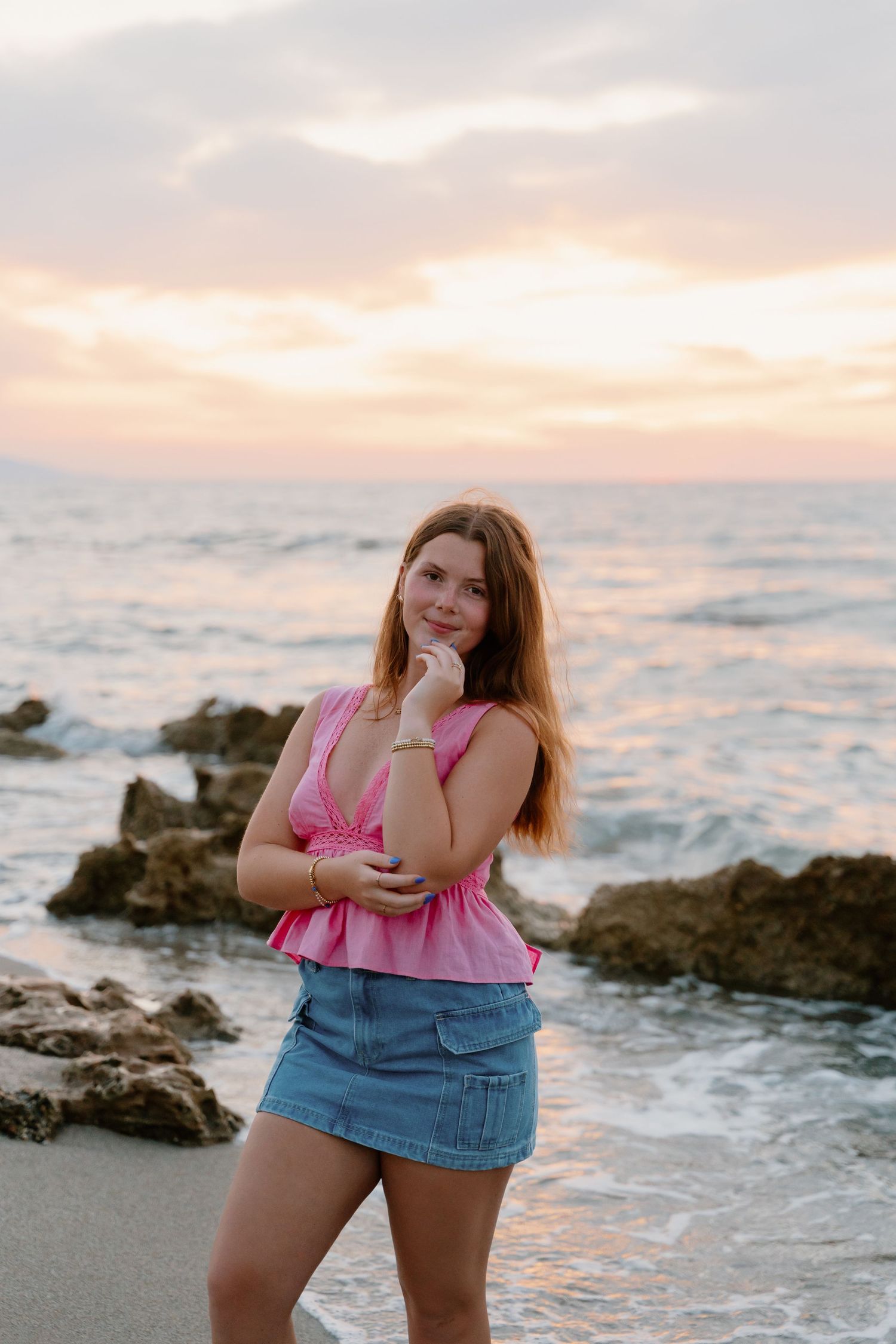 Casual beachside photo with pink top and denim skirt against rocky coastal sunset.