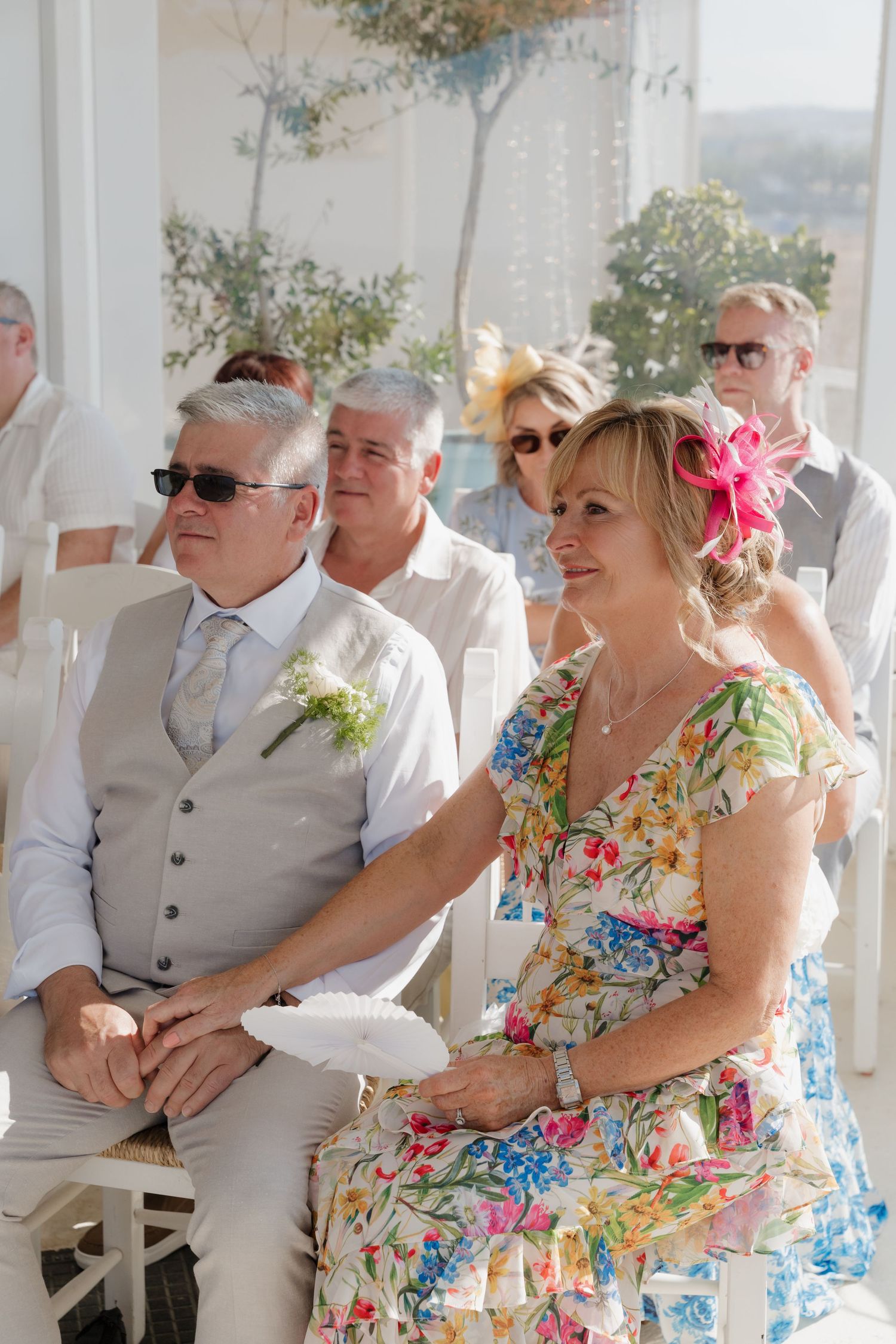 Wedding guests in light summer attire seated at an outdoor venue with floral decorations and sunlit backdrop.