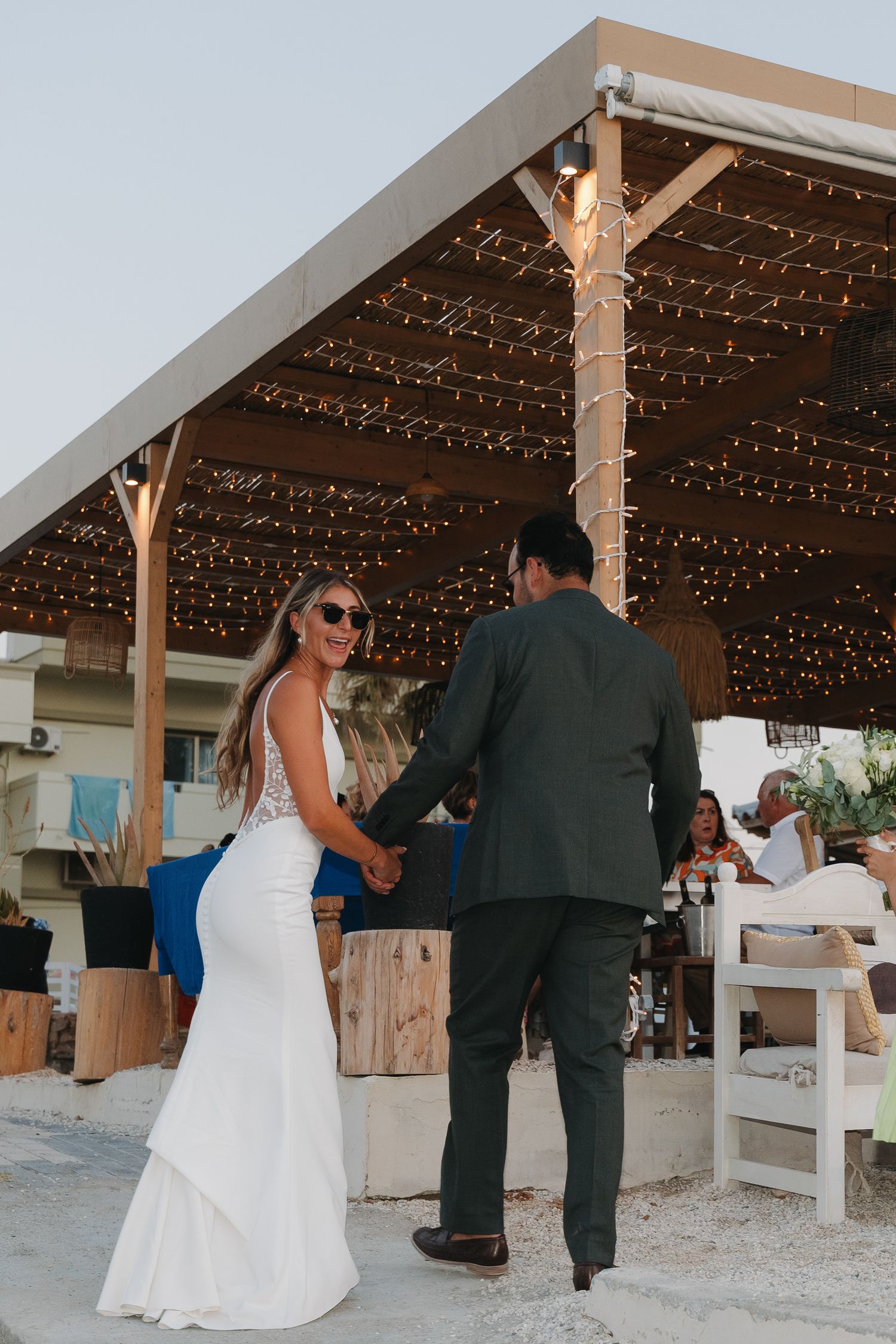 Bride in fitted white dress looks back while walking with groom under string-lit reception pavilion.