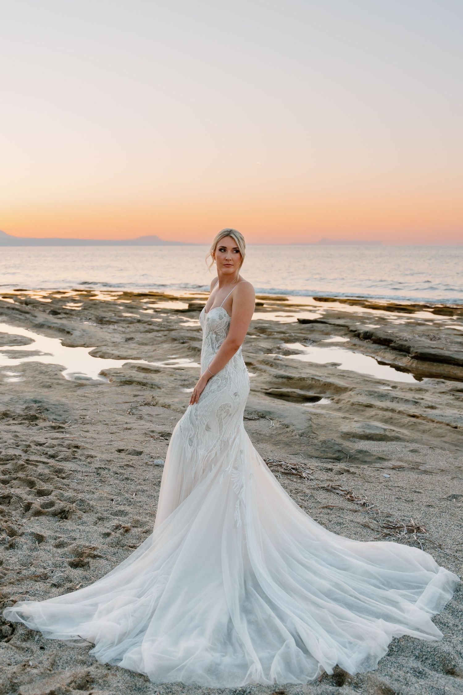 Dramatic bridal portrait with flowing lace gown against sunset coastal backdrop.
