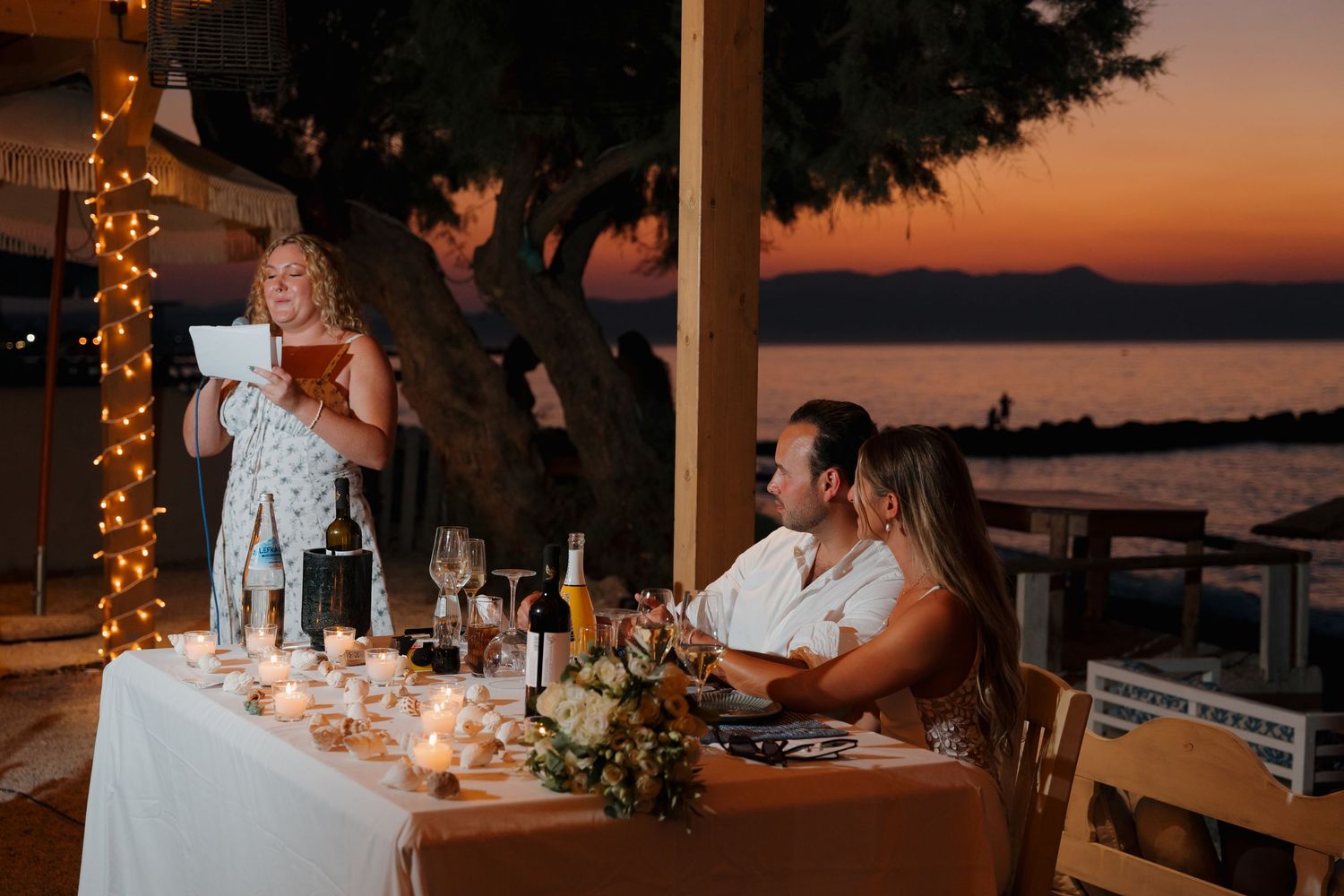 Wedding reception dinner table at sunset with string lights and guests listening to a toast by candlelight.