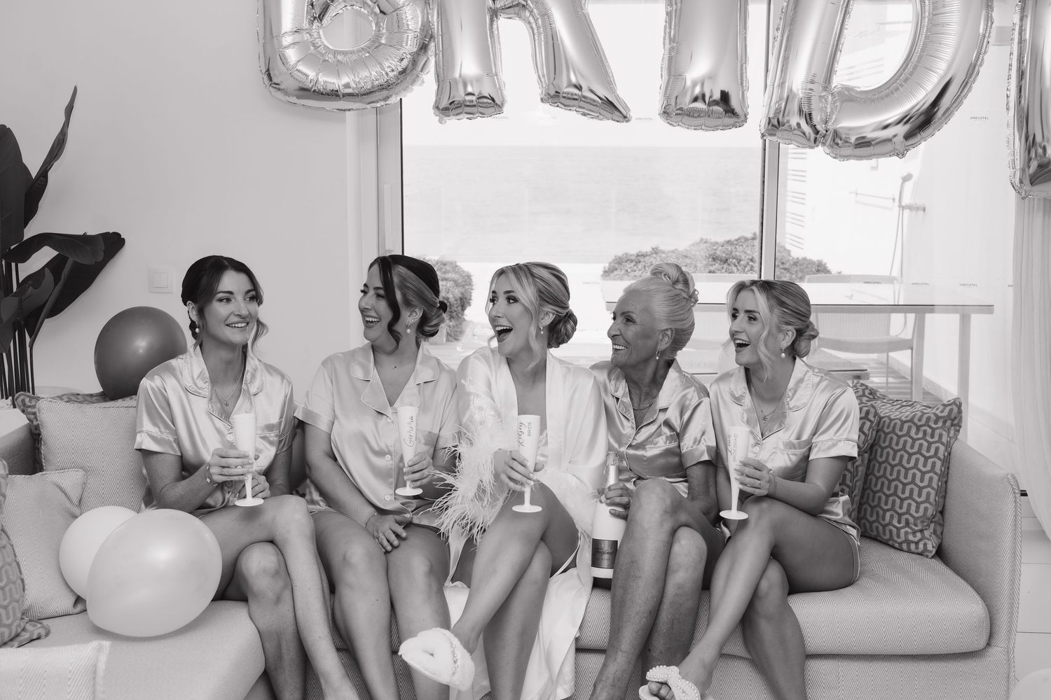 Group of bridesmaids in white robes laughing together on a couch with silver BRIDE balloons in background.