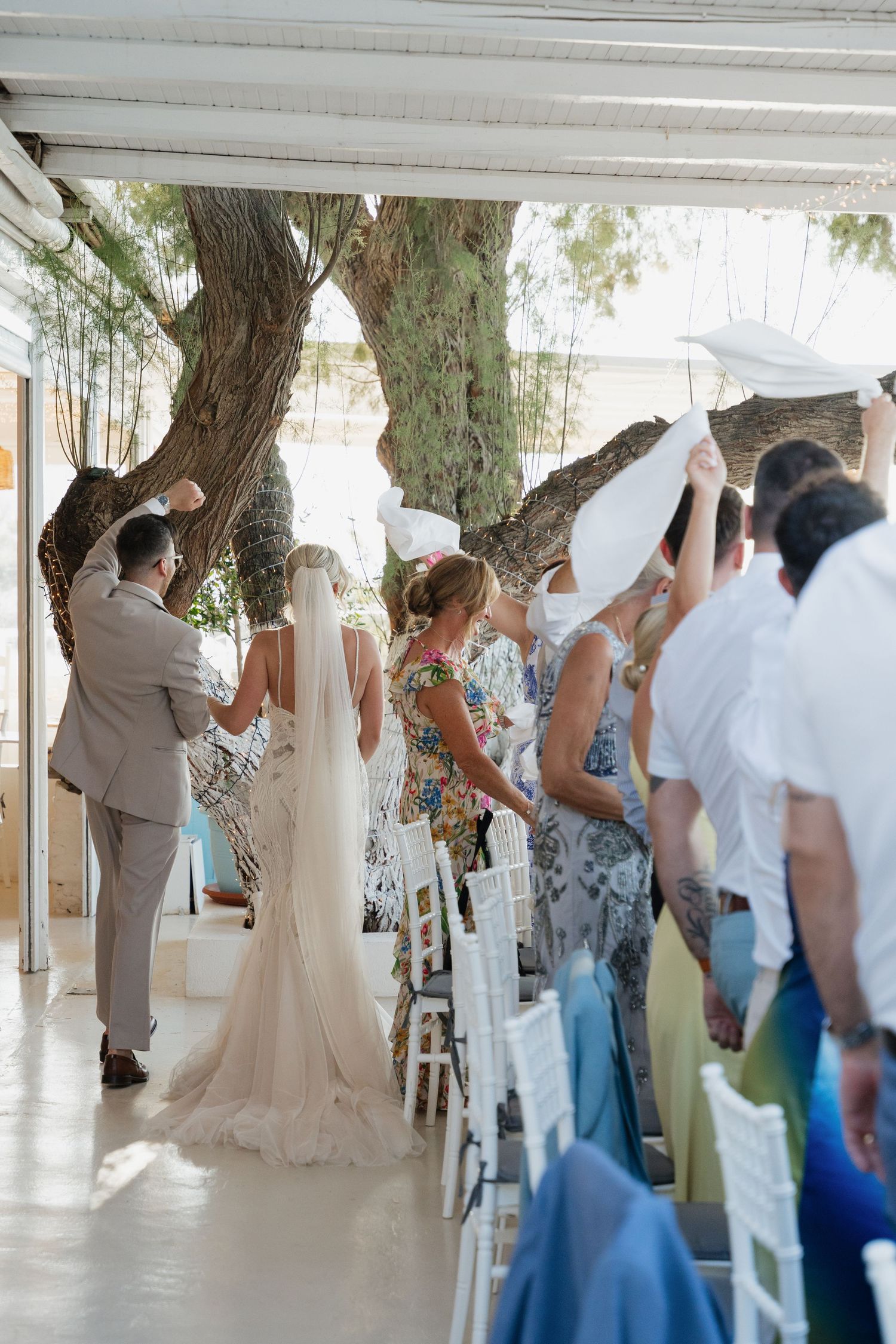 Wedding ceremony guests seated in white chairs under a covered outdoor venue with natural lighting.