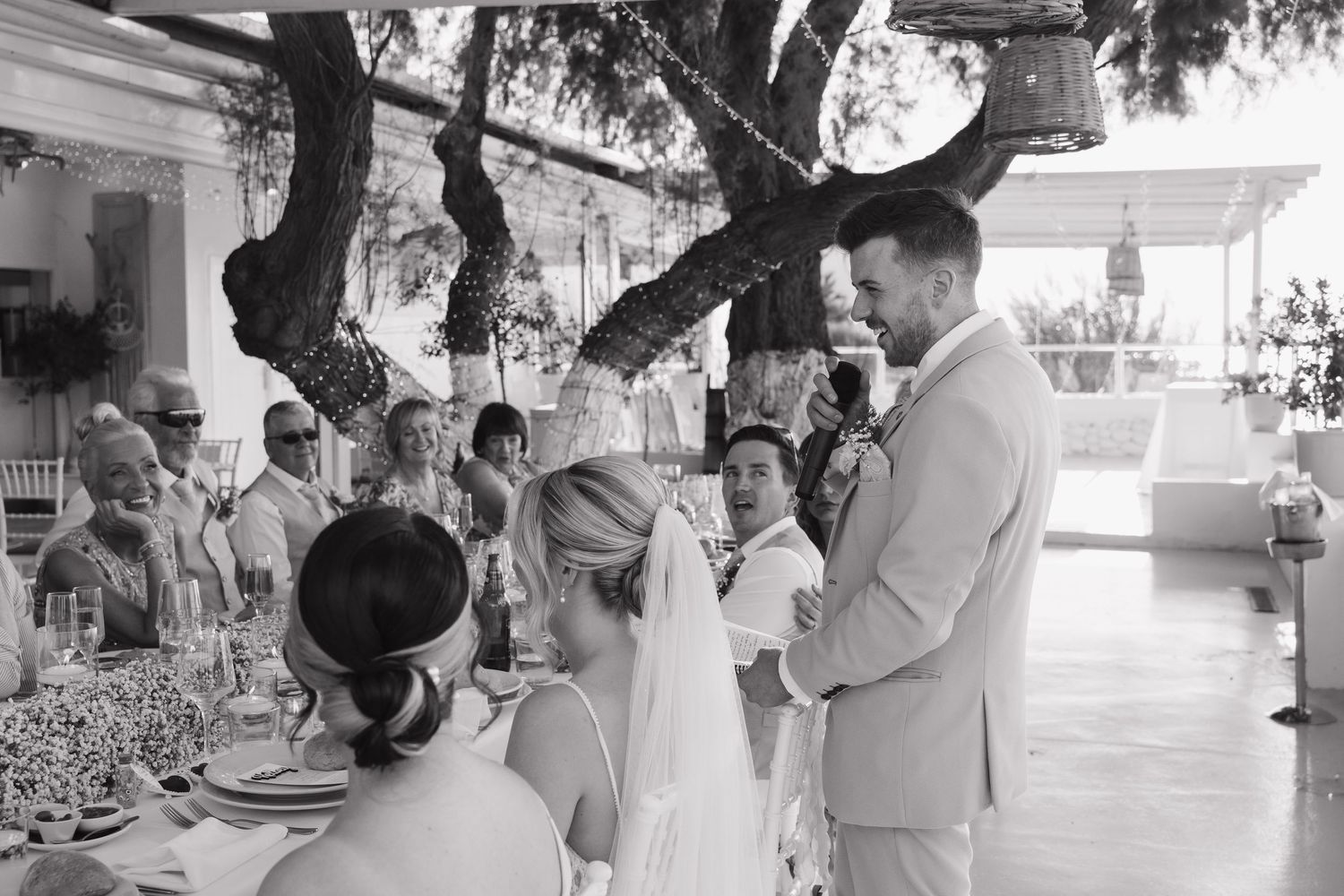 Black and white photo of wedding reception gathering under trees with tables and guests.