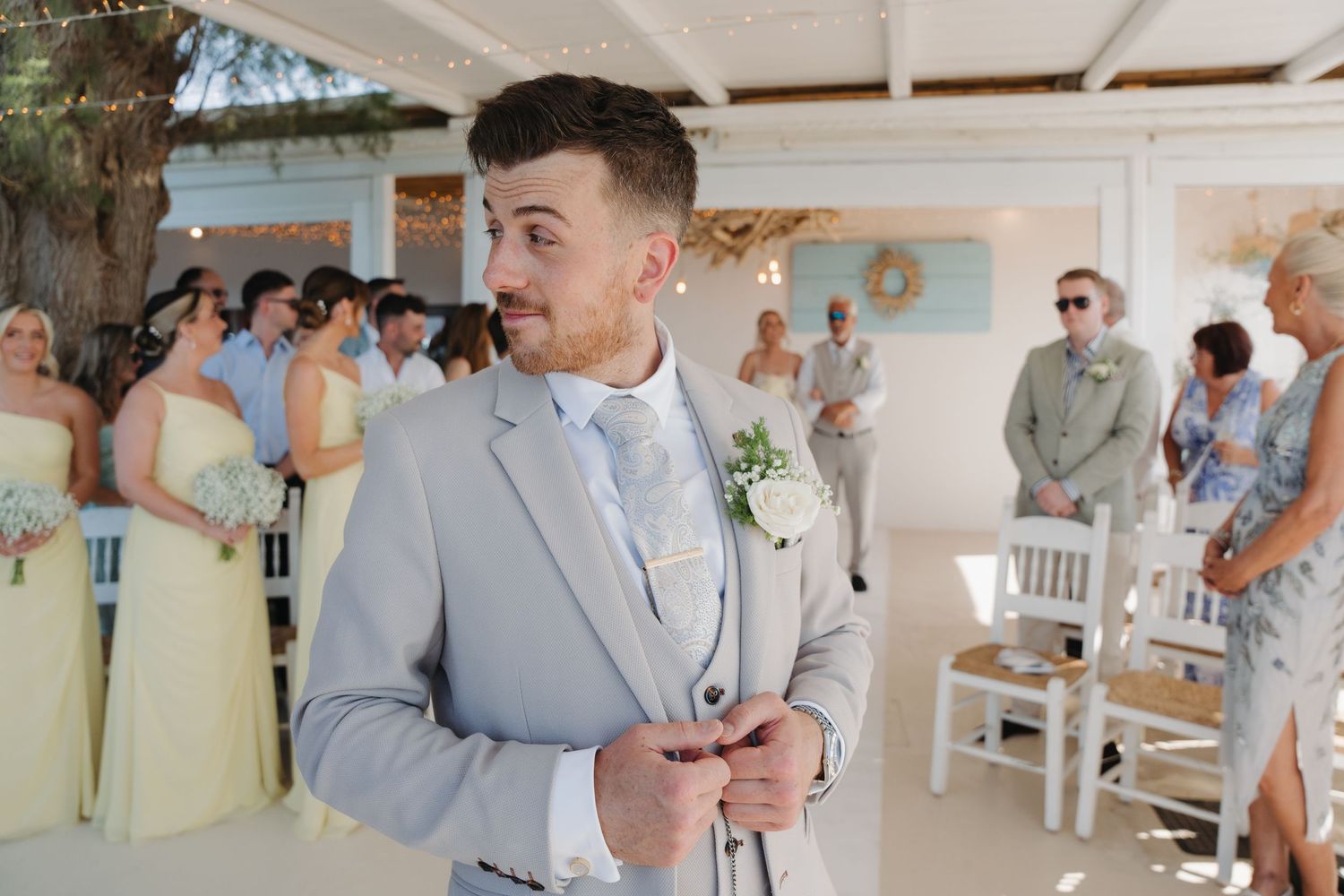 Groom in light gray suit adjusting jacket during outdoor beach wedding ceremony.