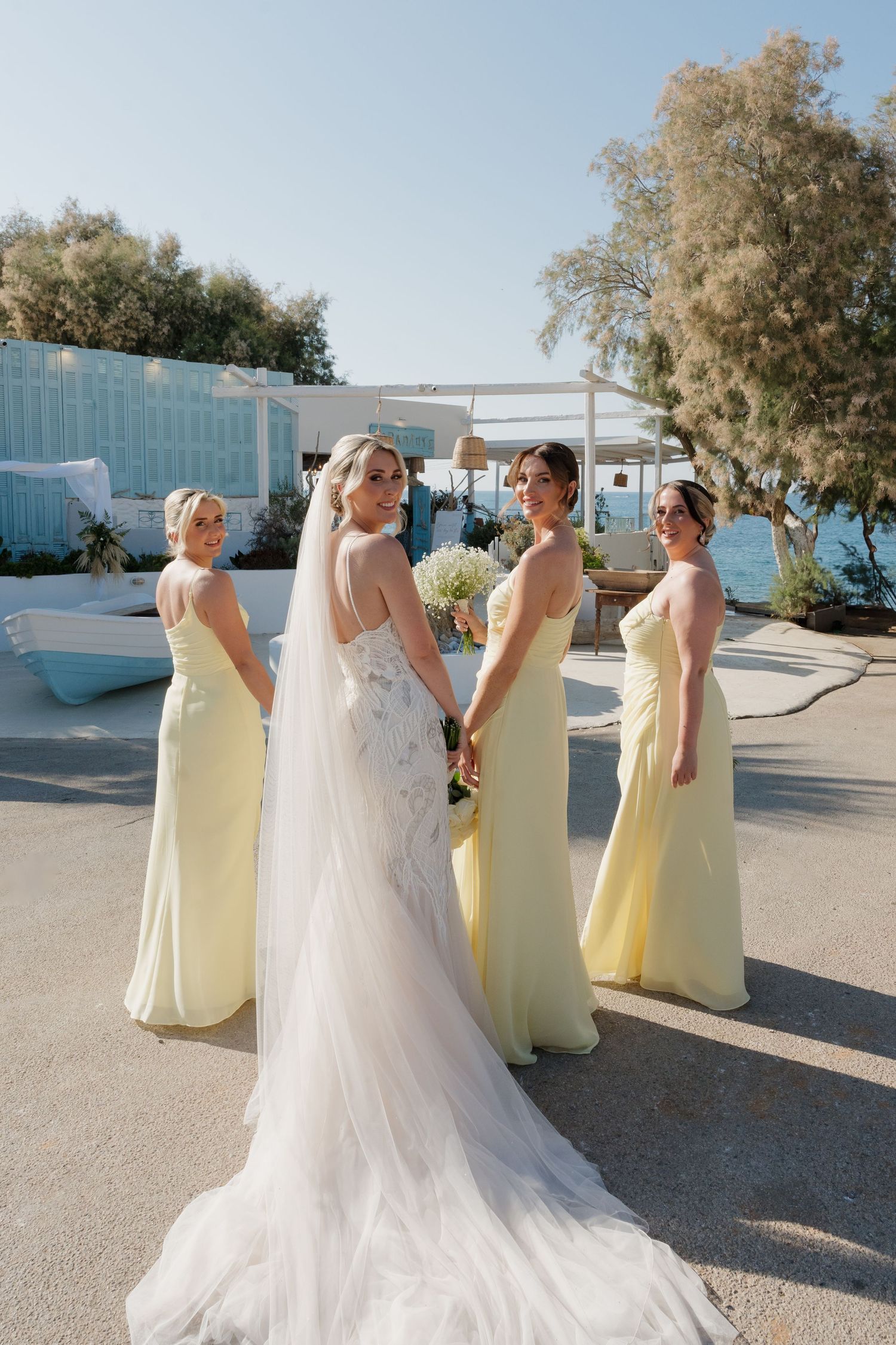 Bridesmaids in pale yellow dresses pose with bride in flowing white gown against seaside backdrop.