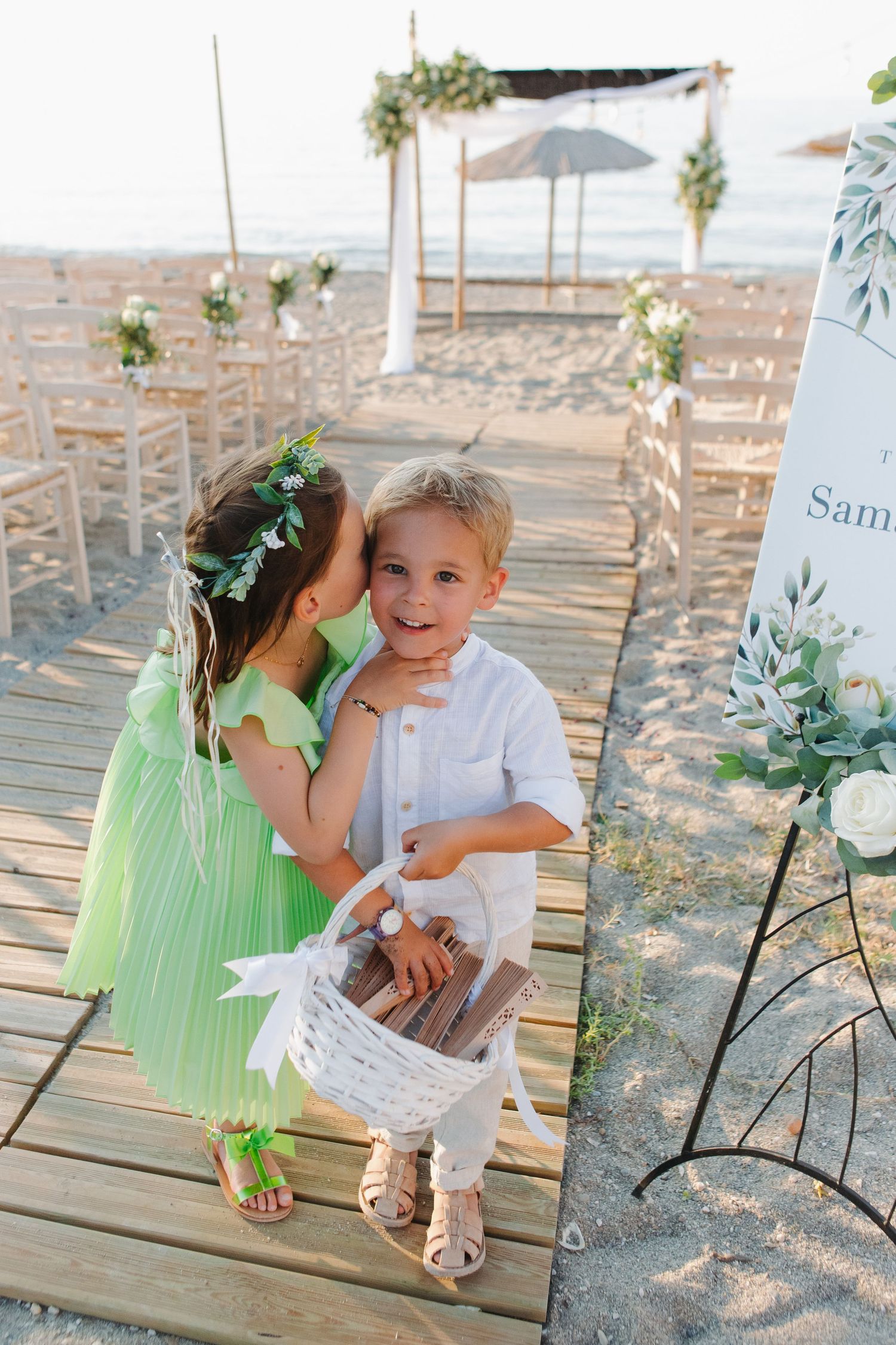 Two young flower children share a sweet moment on wooden walkway at beach wedding ceremony.