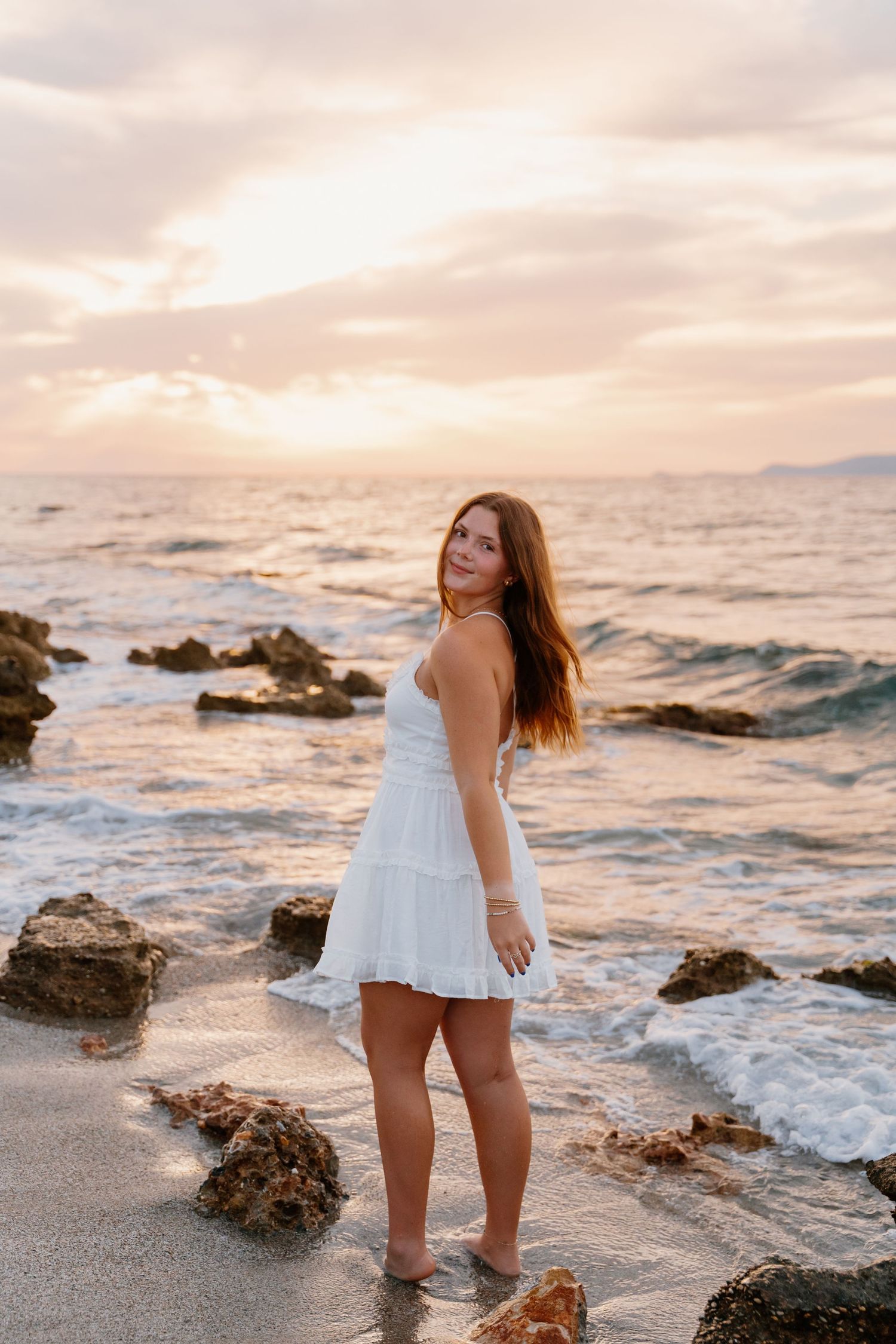 Seaside portrait with white dress against rocky coastline during golden hour.