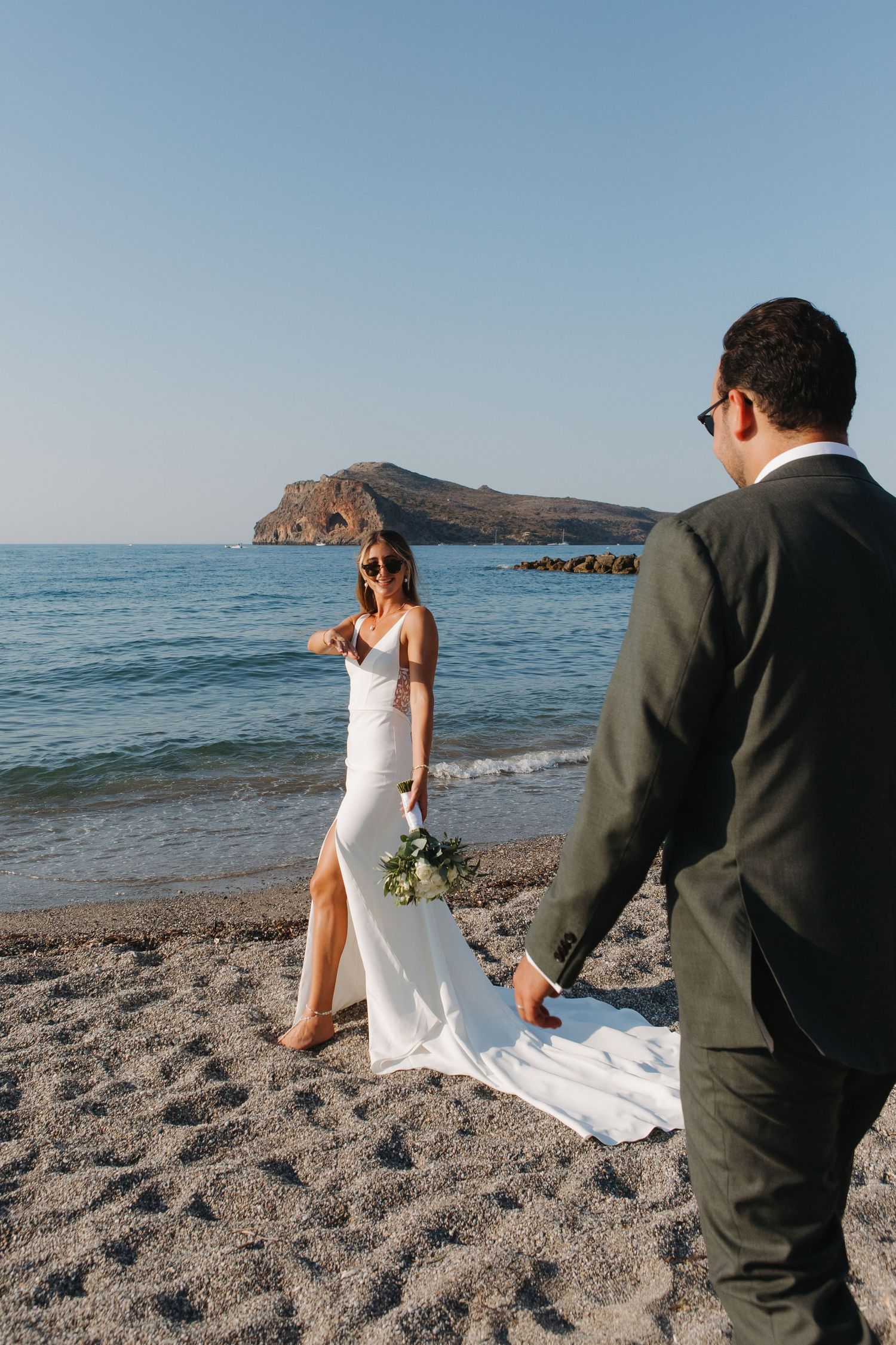 Beach wedding photoshoot with couple posing by ocean with rocky coastline in background.