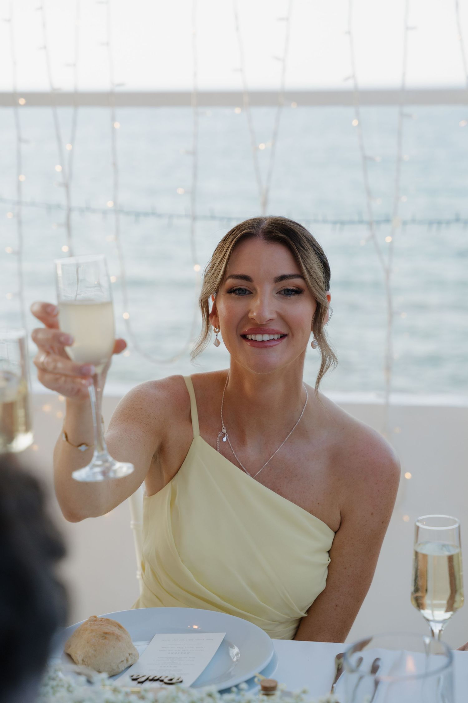 Wedding guest in yellow dress raises champagne glass for toast with ocean view in background.