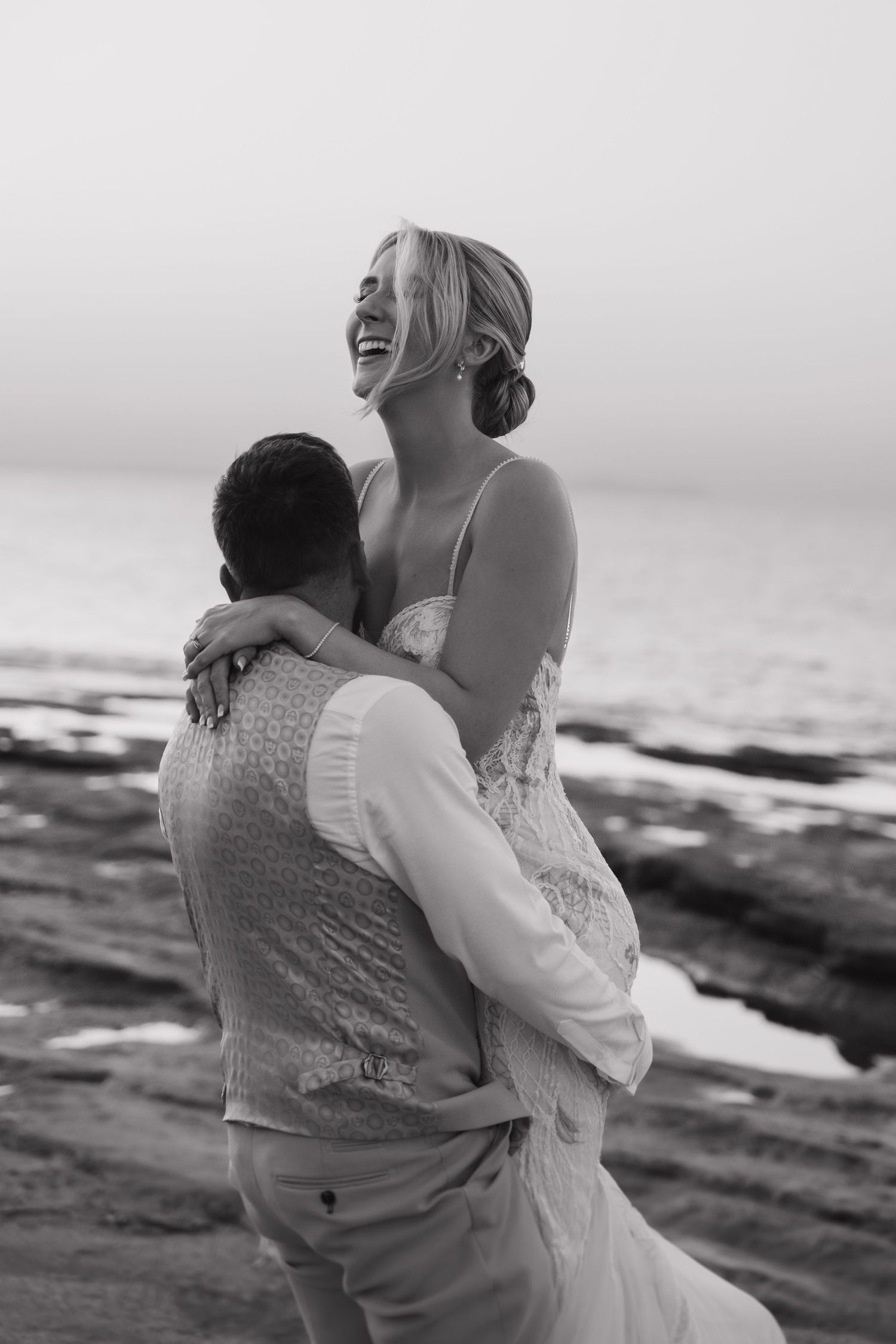 Black and white candid photo of happy couple embracing on beach rocks at sunset.