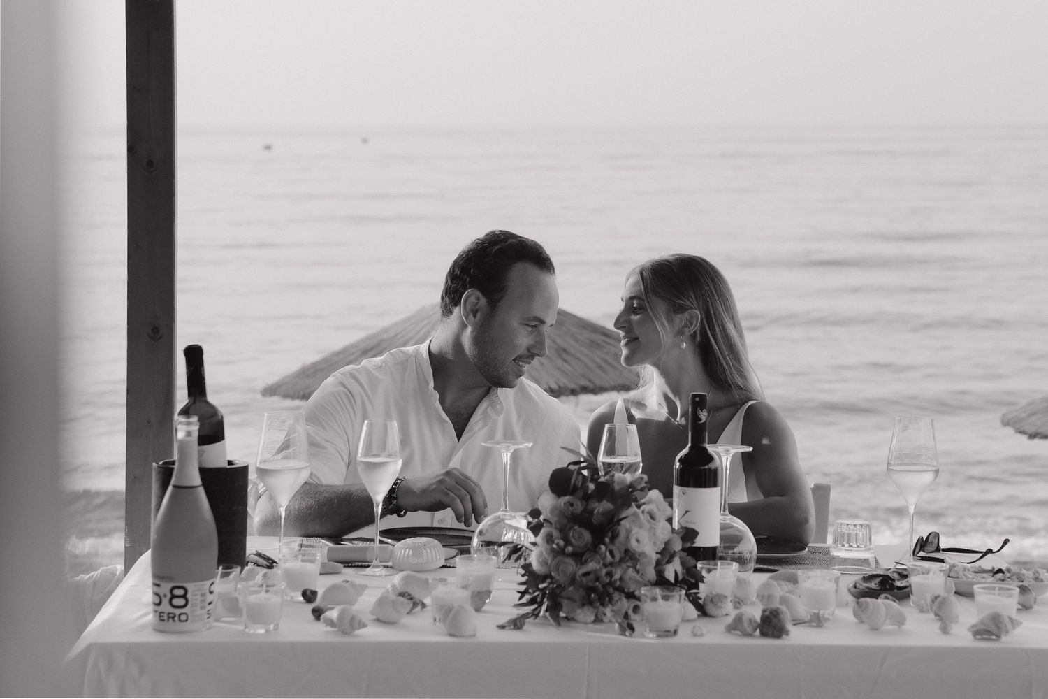 An intimate black and white dinner scene of a couple sharing a moment at a seaside table.