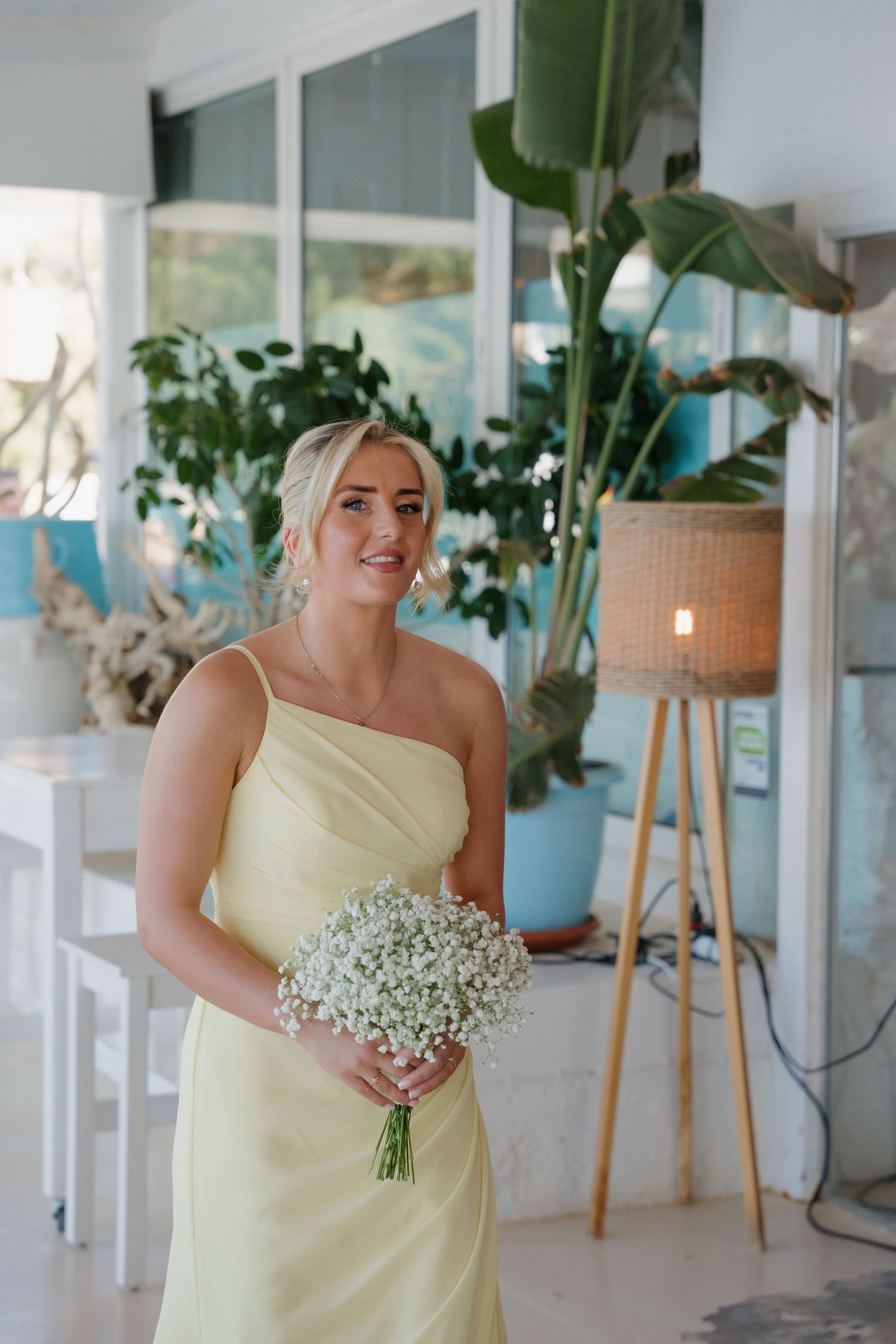 Bridesmaid in pale yellow dress holds baby's breath bouquet in bright indoor setting.
