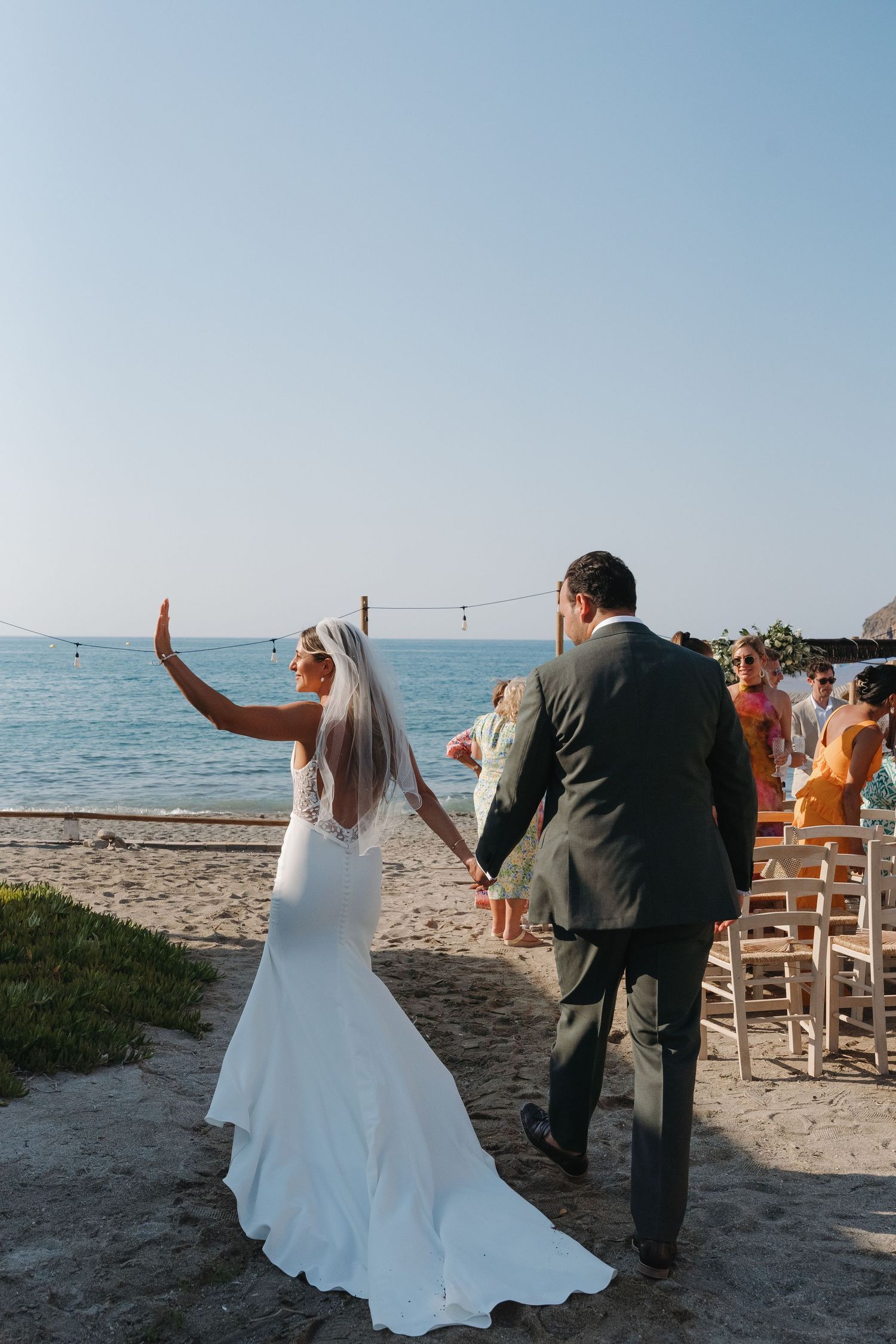 A beach wedding celebration with guests seated on wooden chairs by the ocean under blue skies.