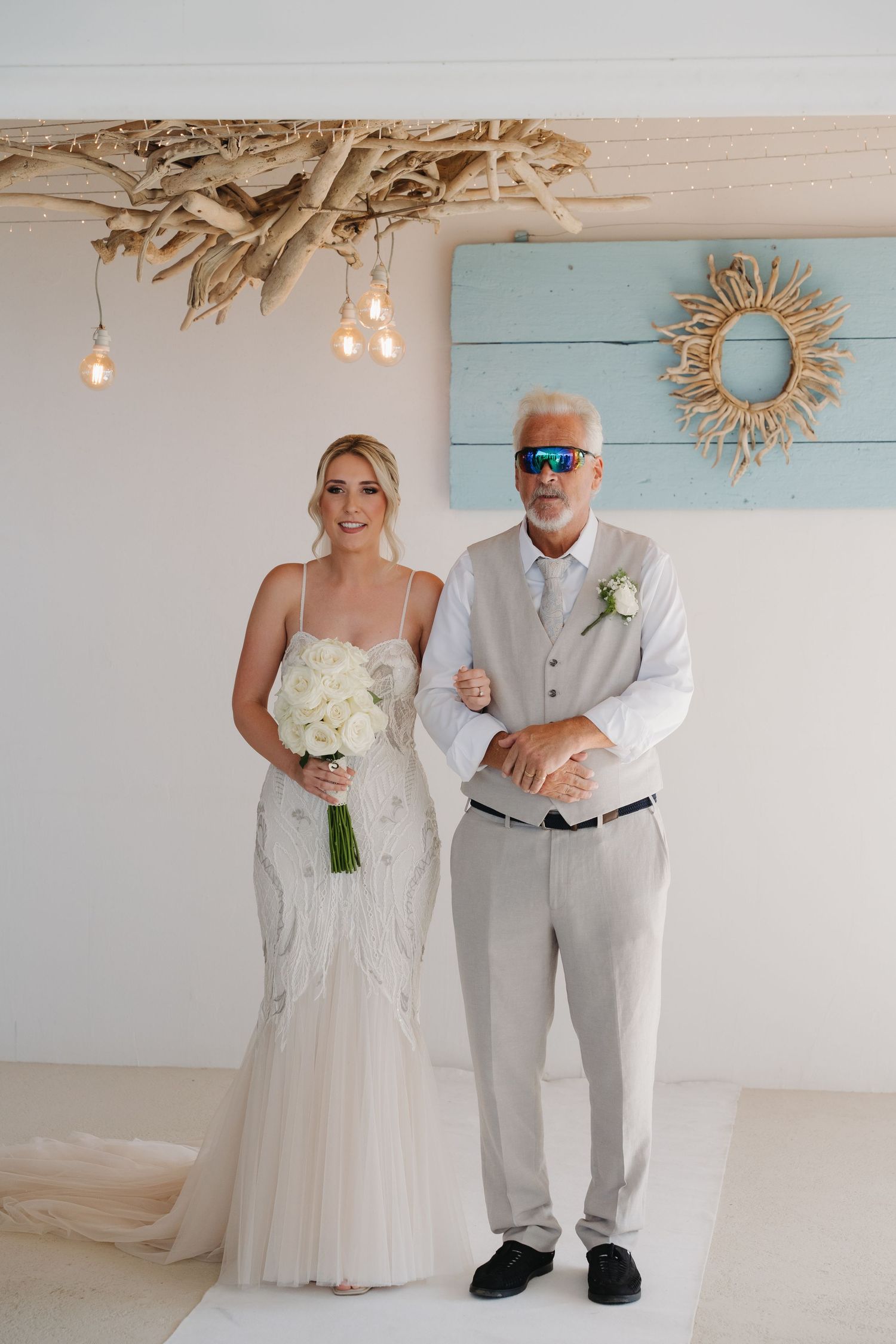 Wedding couple poses under driftwood chandelier in coastal venue setting.