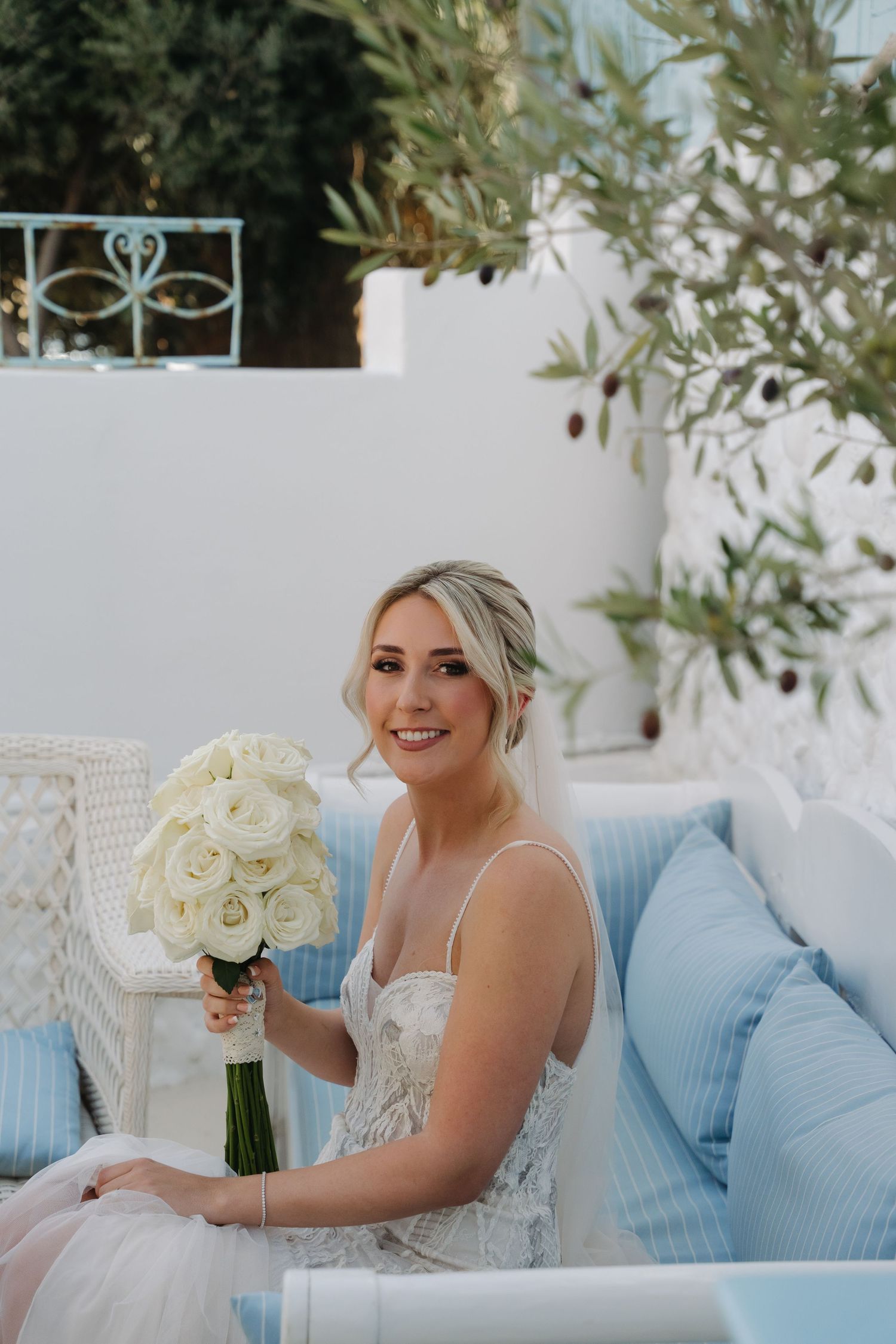 A person in a white wedding dress sits on blue cushions holding a bouquet of white roses.