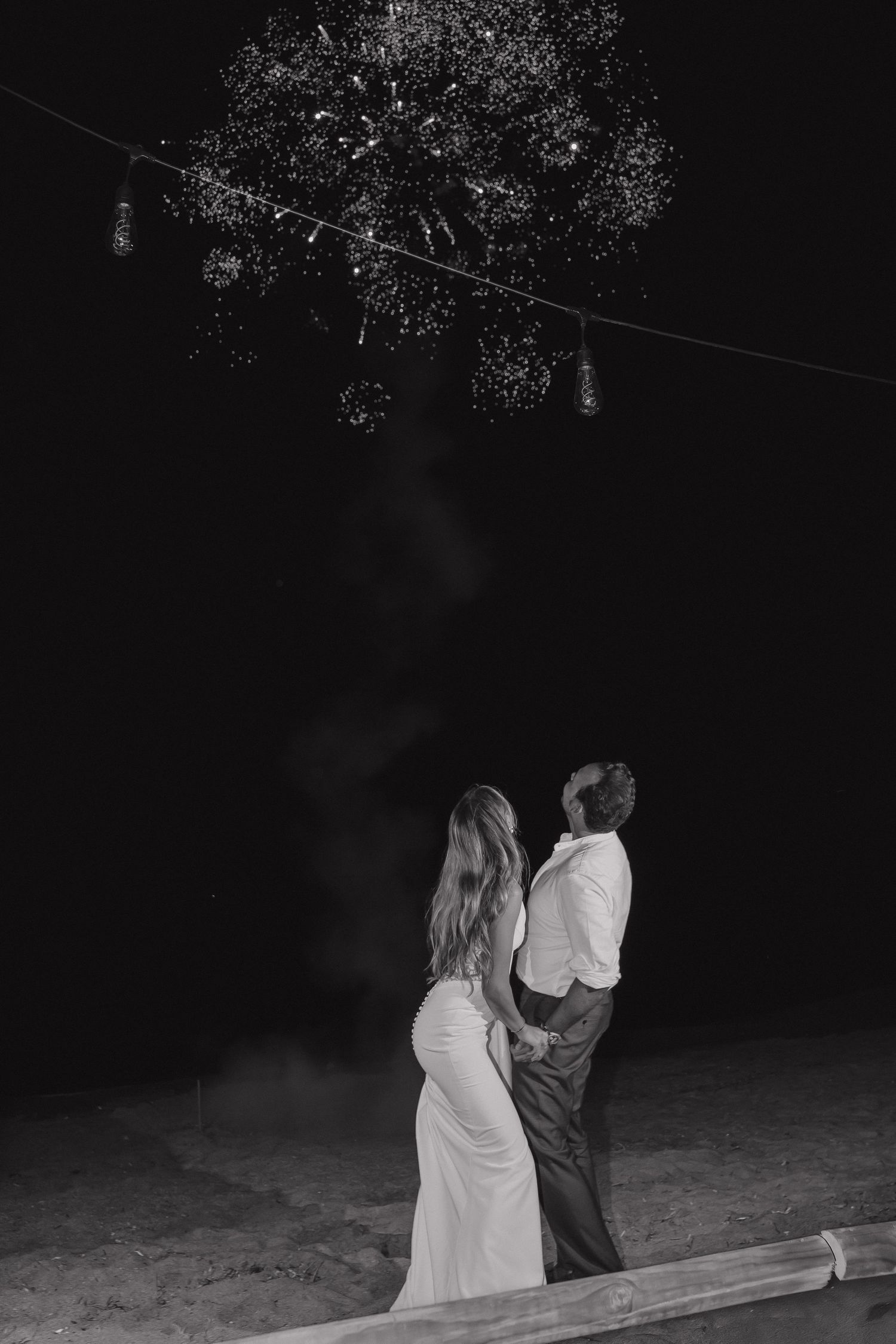 Black and white photo of couple gazing at string lights during nighttime celebration.
