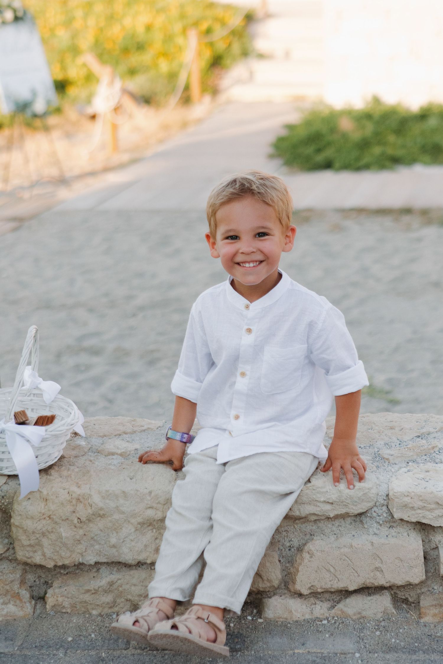Young ring bearer in white shirt and khaki pants sits on stone wall during beach wedding.
