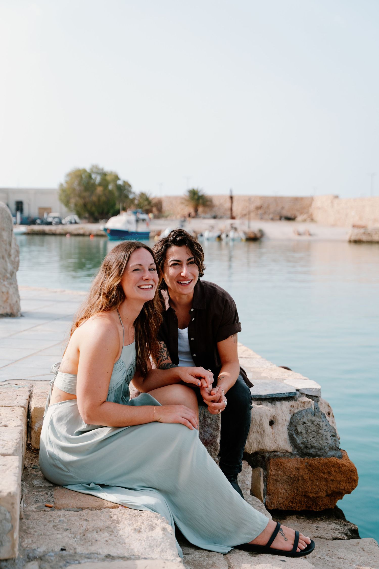Two people share a joyful moment while sitting on stone steps in Rethymno, Crete near turquoise water at a Mediterranean harbor.
