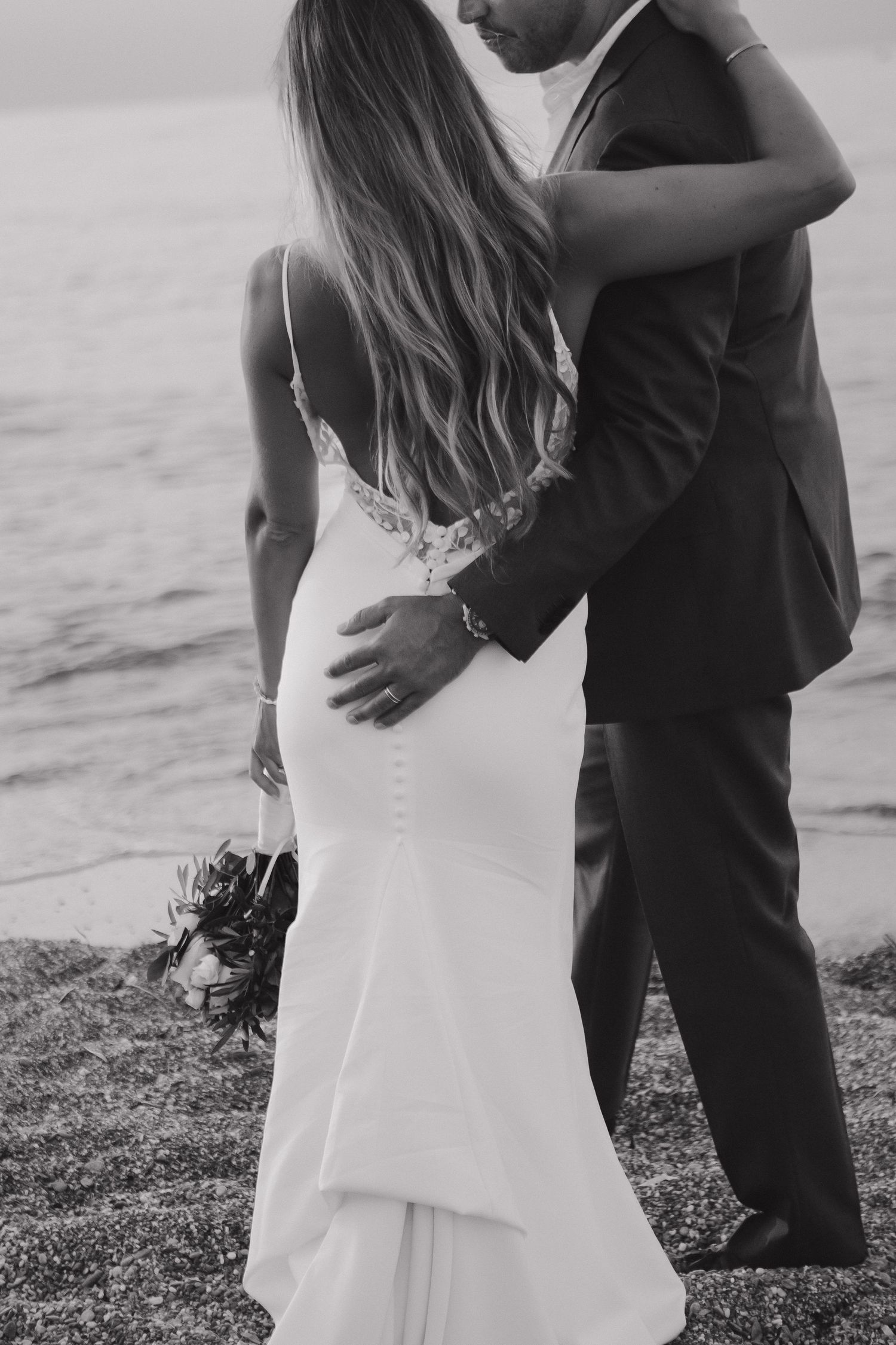 A romantic black and white beach wedding photo showing a couple embracing with the bride in a fitted lace gown.