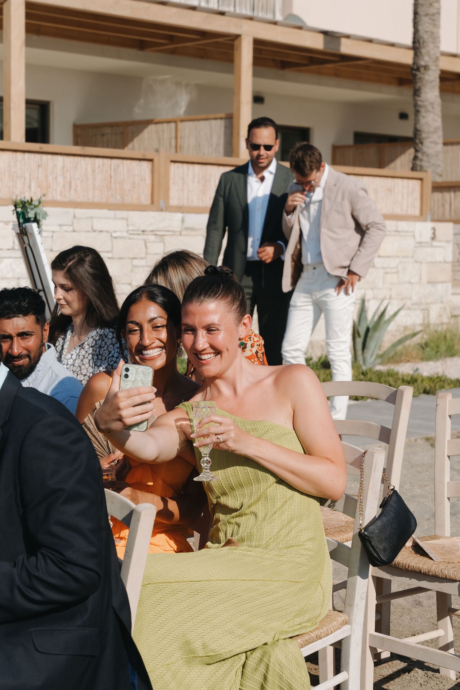 Guests at an outdoor party laugh while looking at phones together on a sunny patio.