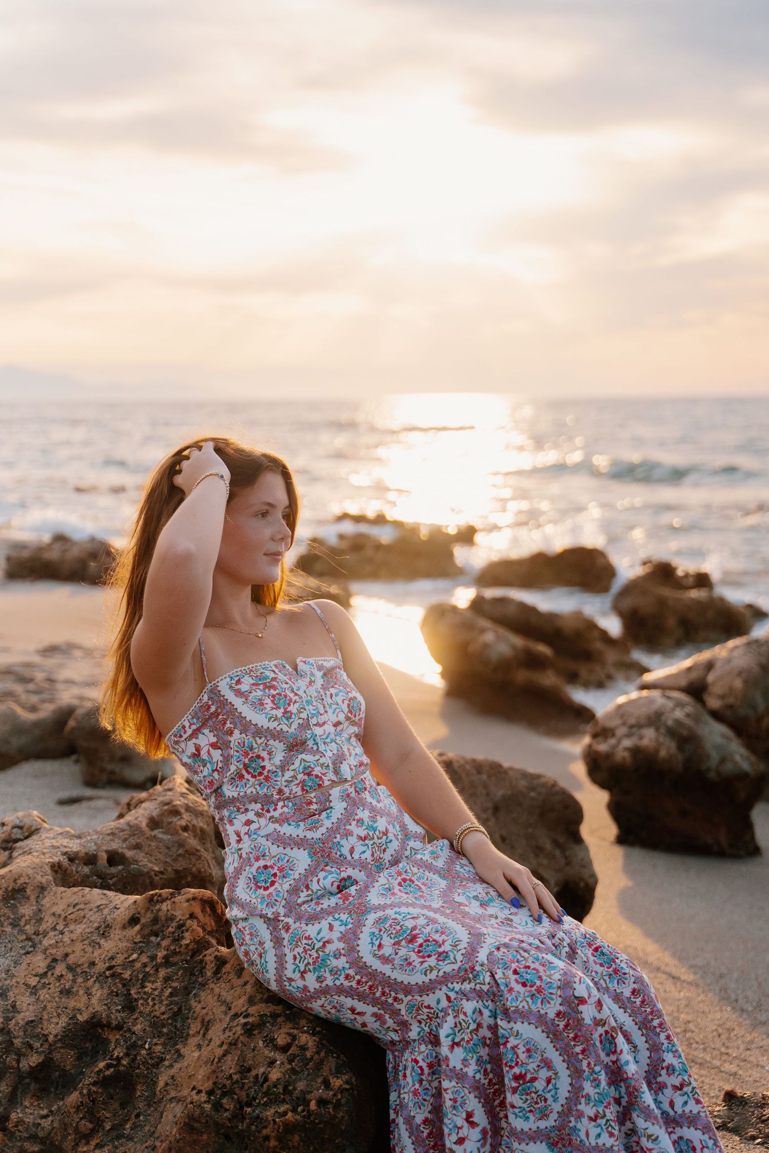Woman in patterned dress posing on rocky beach during golden hour sunset.