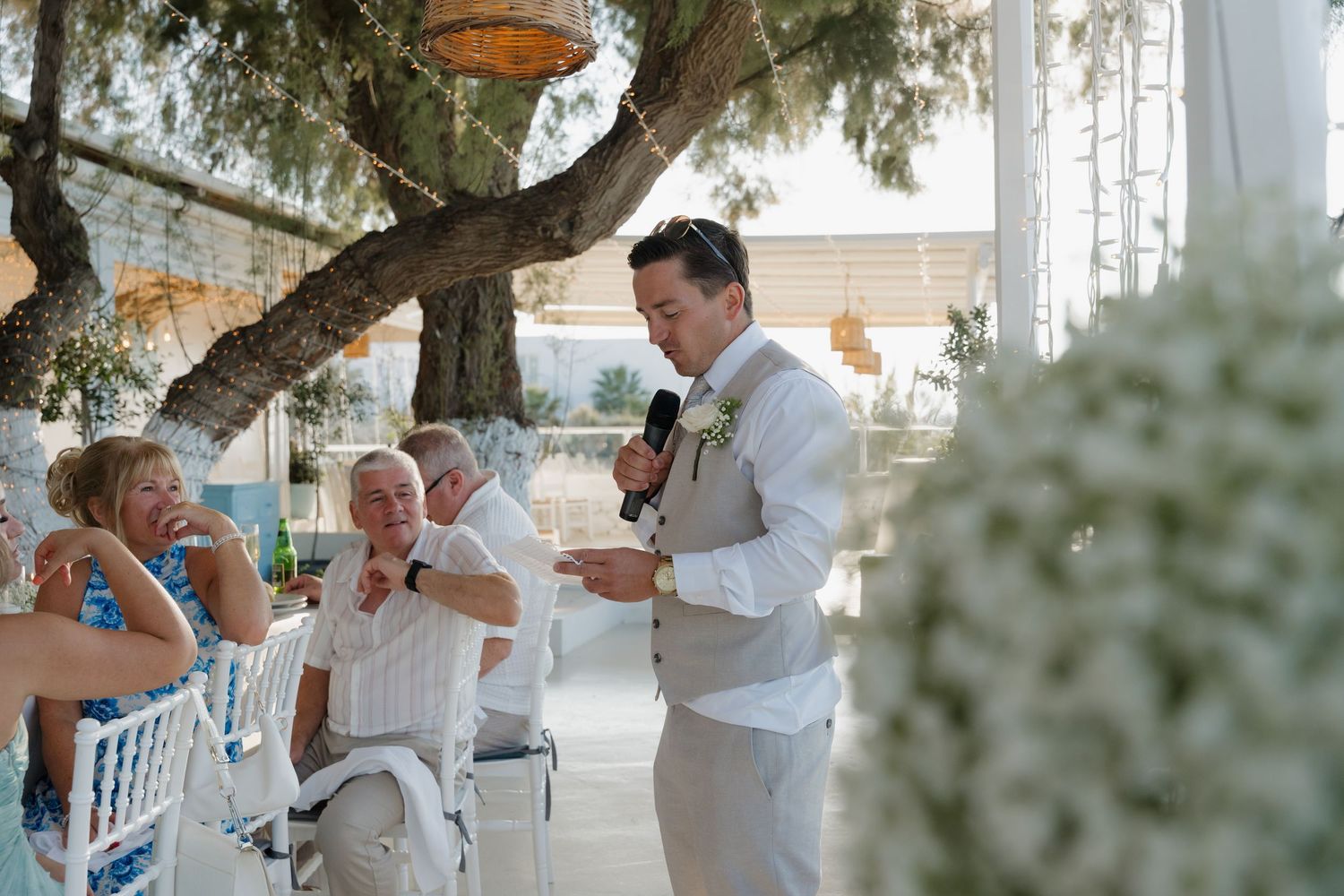 People gathered outdoors for a celebration event with a speaker in white under a tree with hanging decorations.