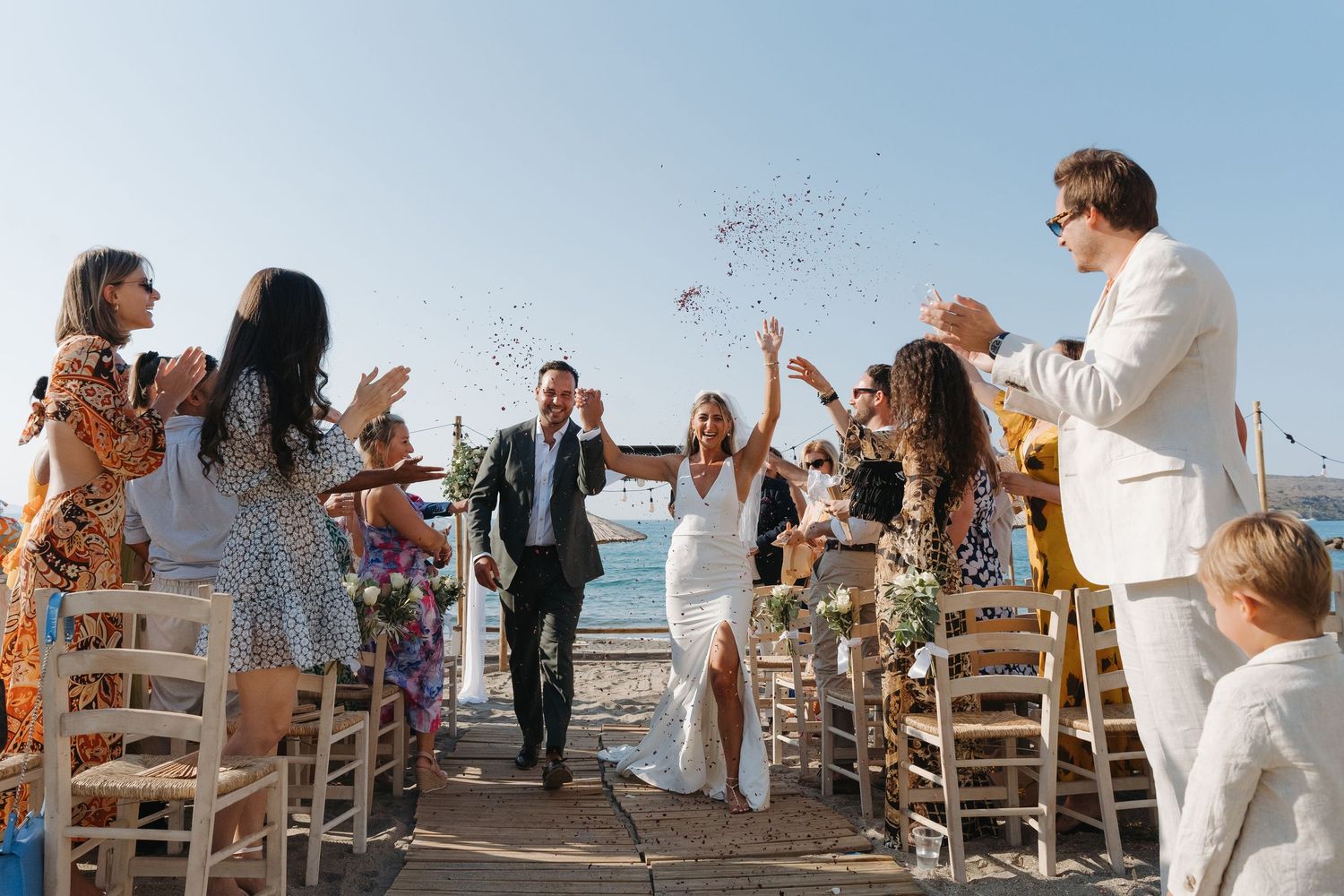 Joyful wedding celebration with guests tossing confetti on a seaside deck.
