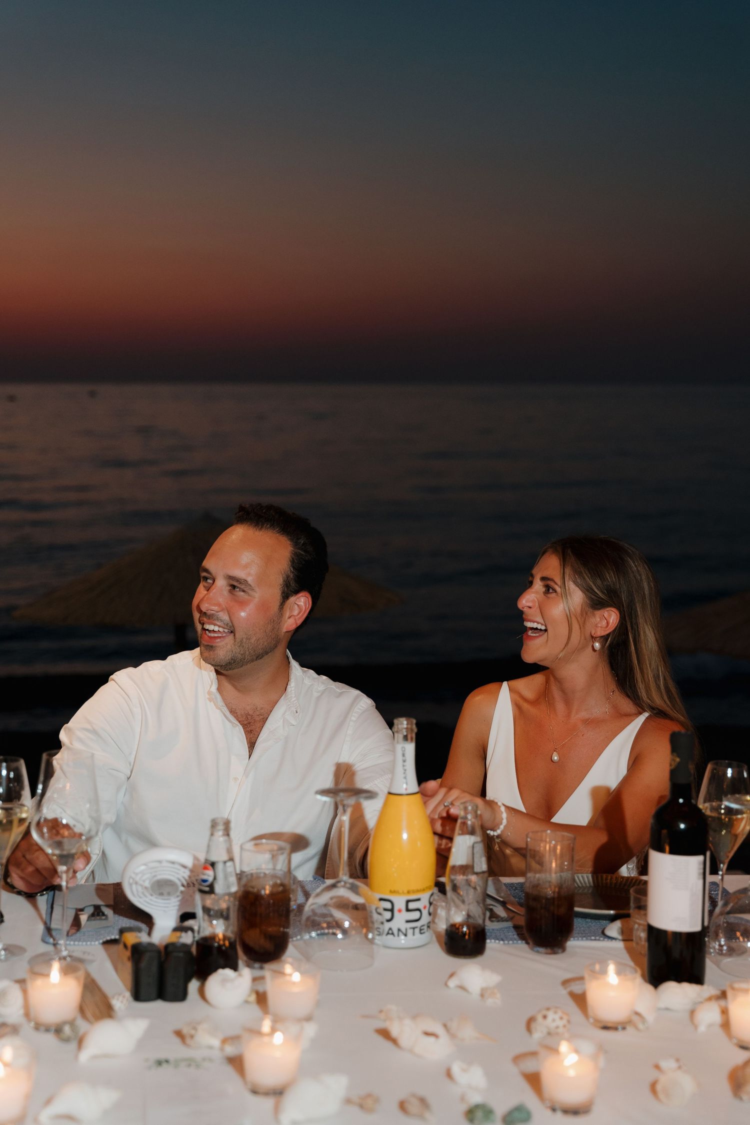 Newlyweds laugh together at candlelit reception dinner table by the ocean.