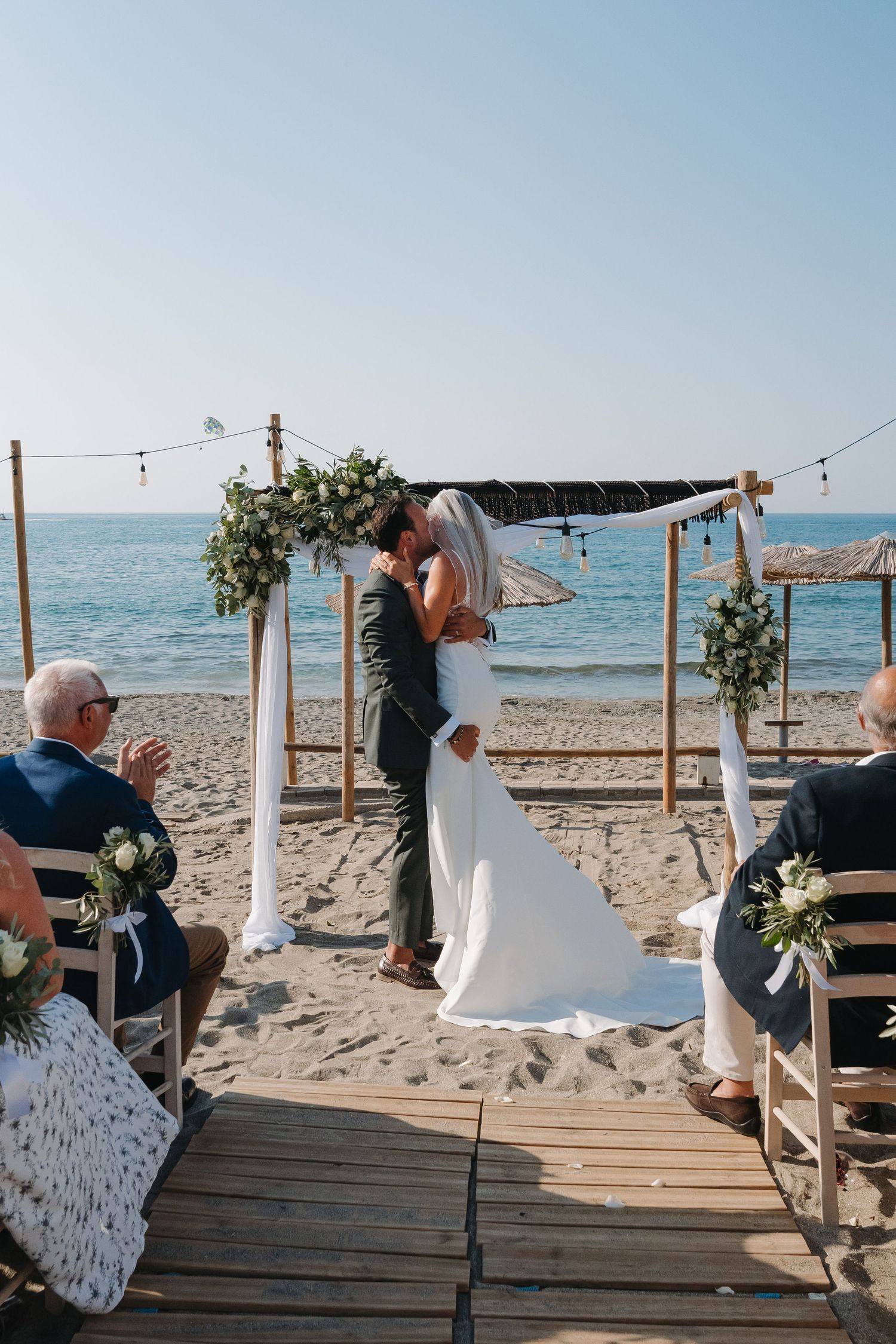 A wooden boardwalk beach wedding setup with string lights and white fabric decorations.