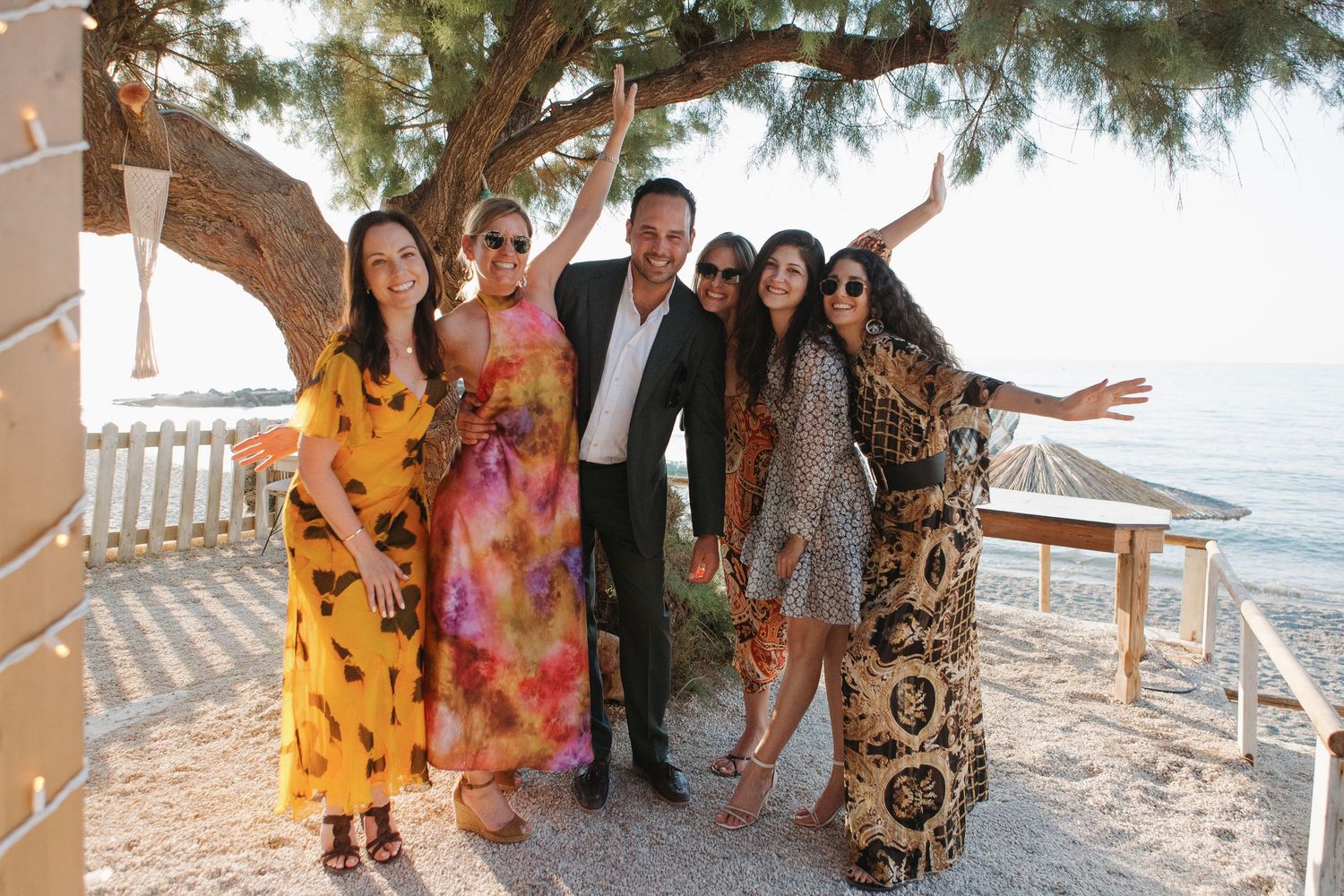 Wedding guests in colorful attire gather for a group photo by the ocean.