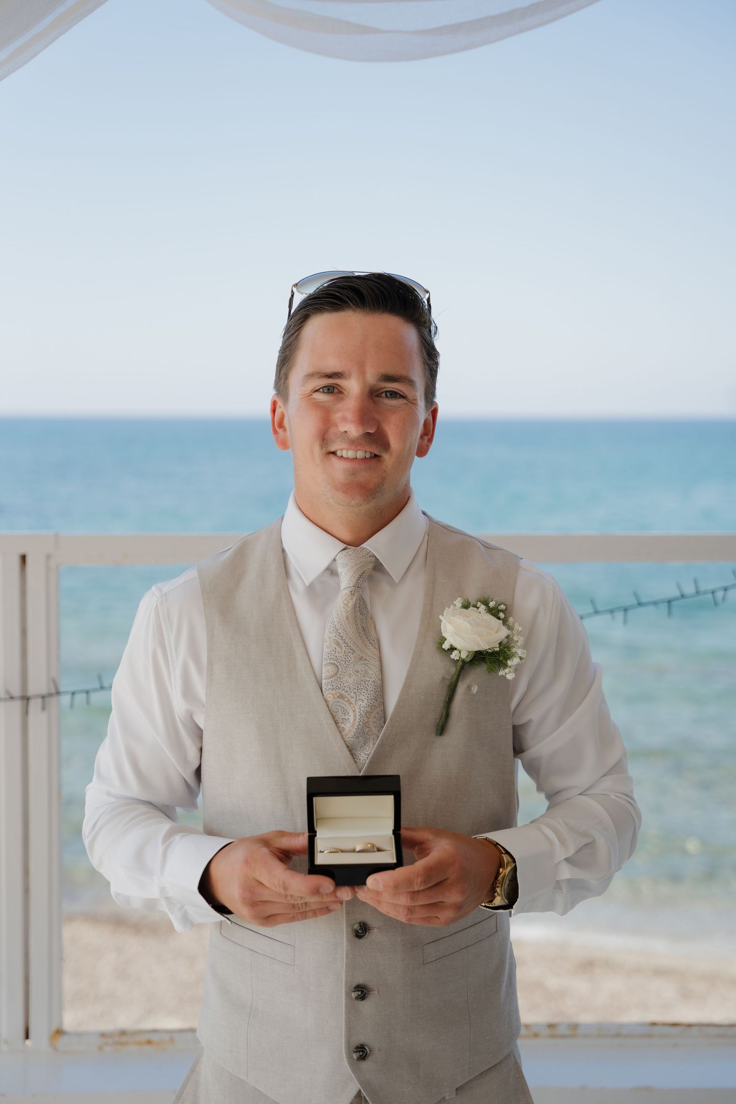 Person in light colored wedding attire holding a ring box against an ocean backdrop.