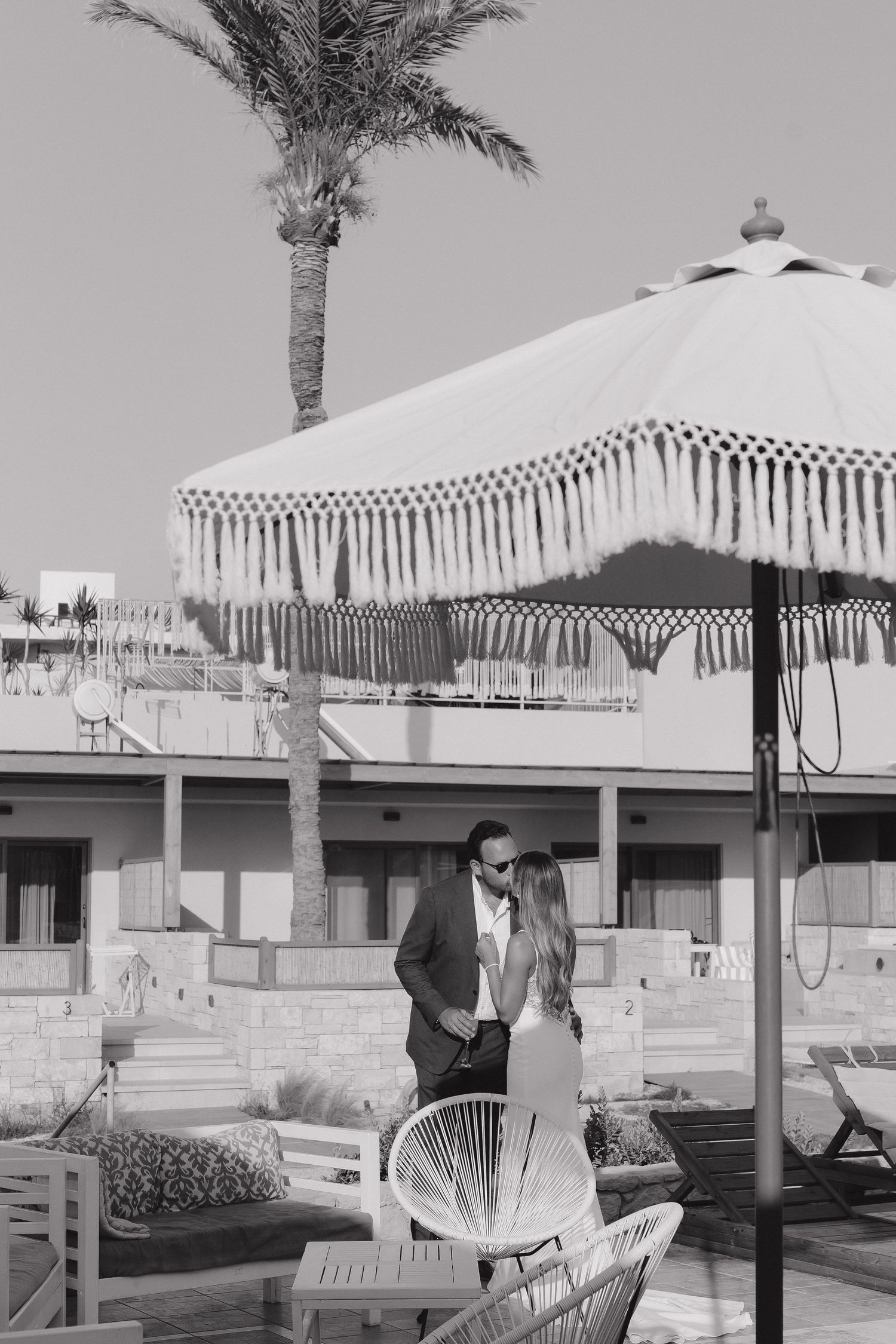 A black and white photo of a beachside venue with a decorative fringed umbrella and palm tree.