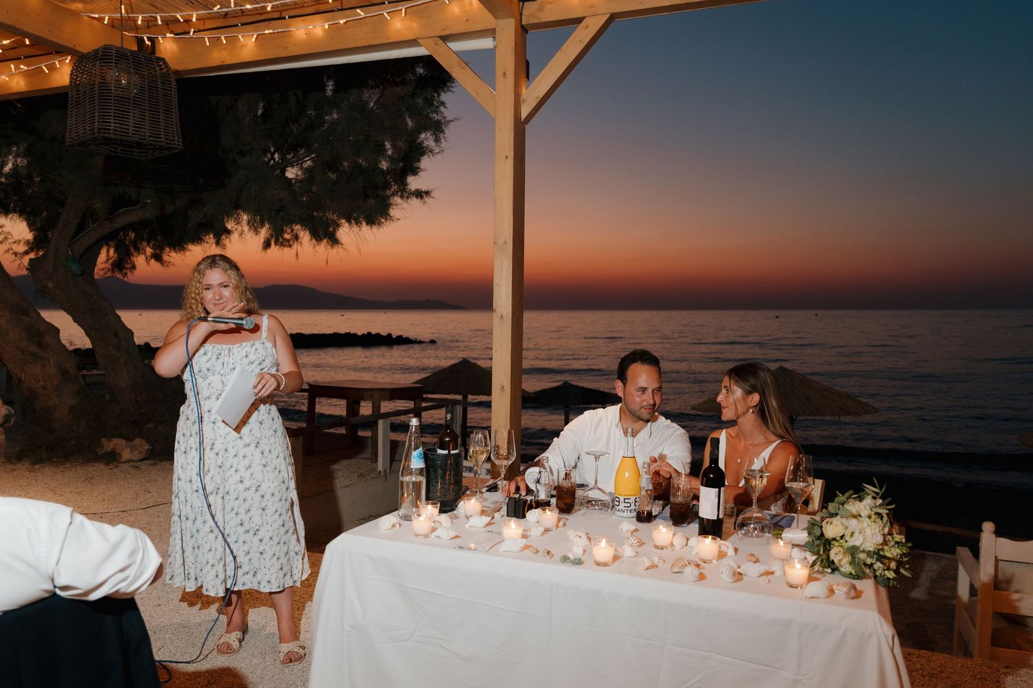 Wedding reception dinner setup with string lights and ocean sunset view.