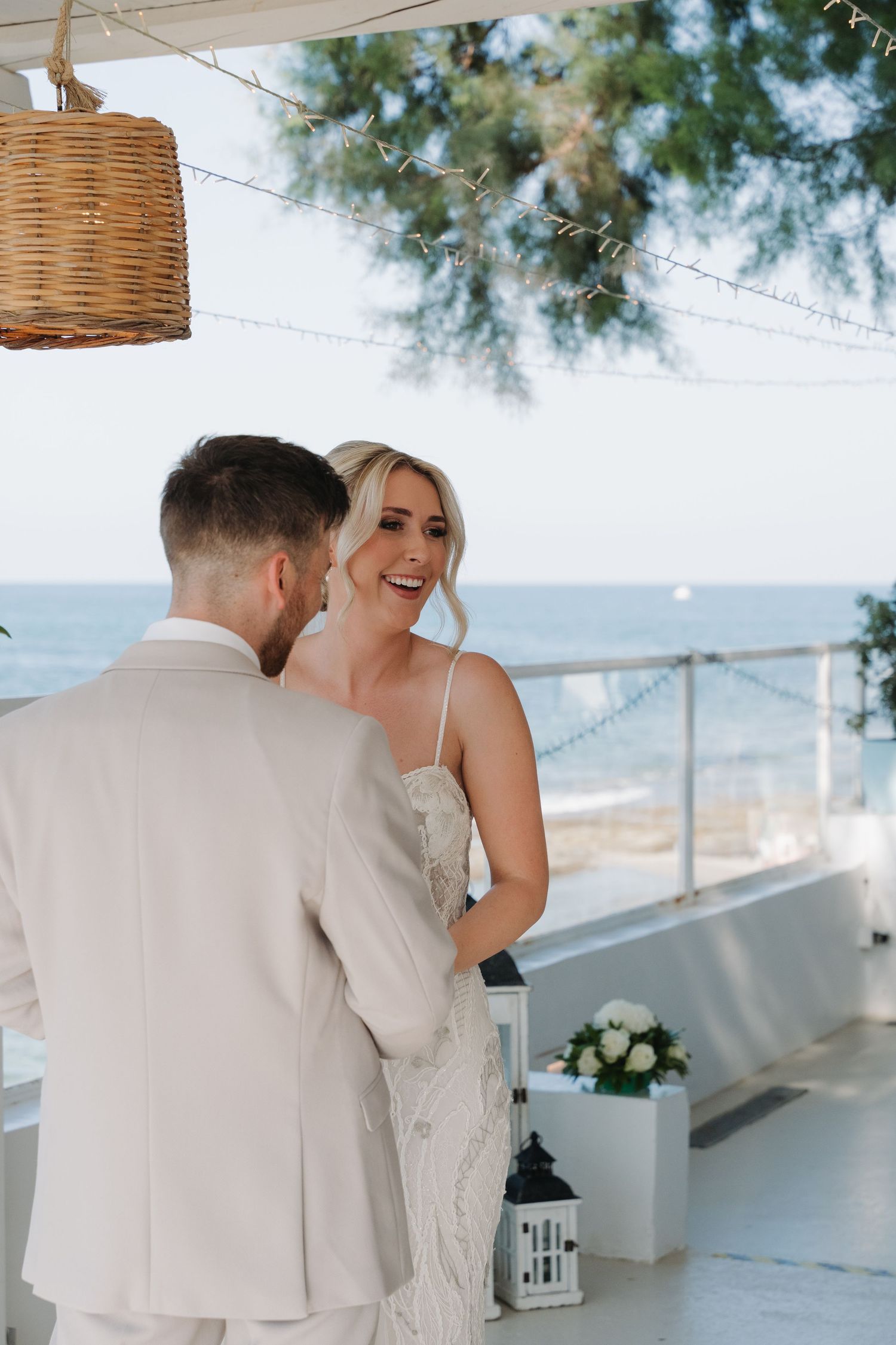 A romantic wedding moment on a beachfront terrace with wicker pendant lights and ocean views.