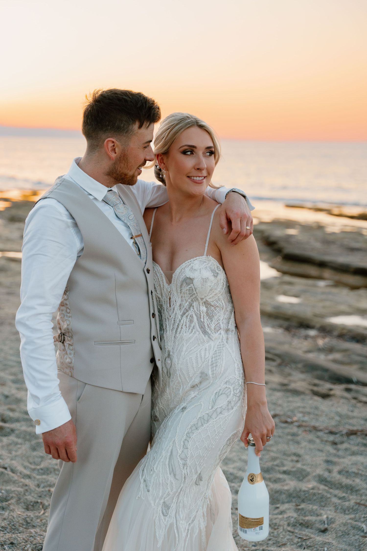 Newlywed couple poses on beach at sunset in elegant white wedding attire.