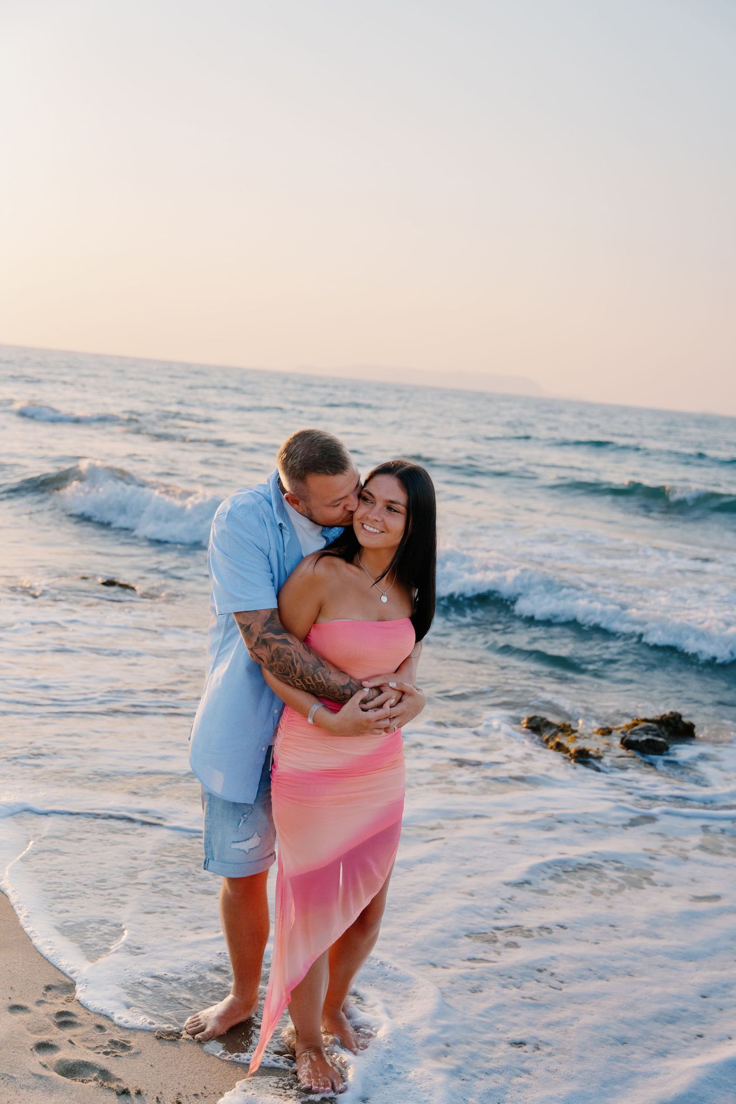 A couple embraces on the beach at sunset with waves crashing behind them.