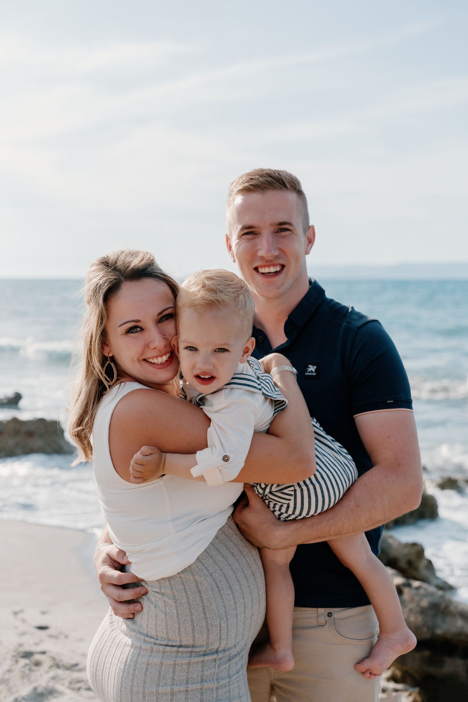 Loving family portrait on the beach with ocean waves and rocks in the background.