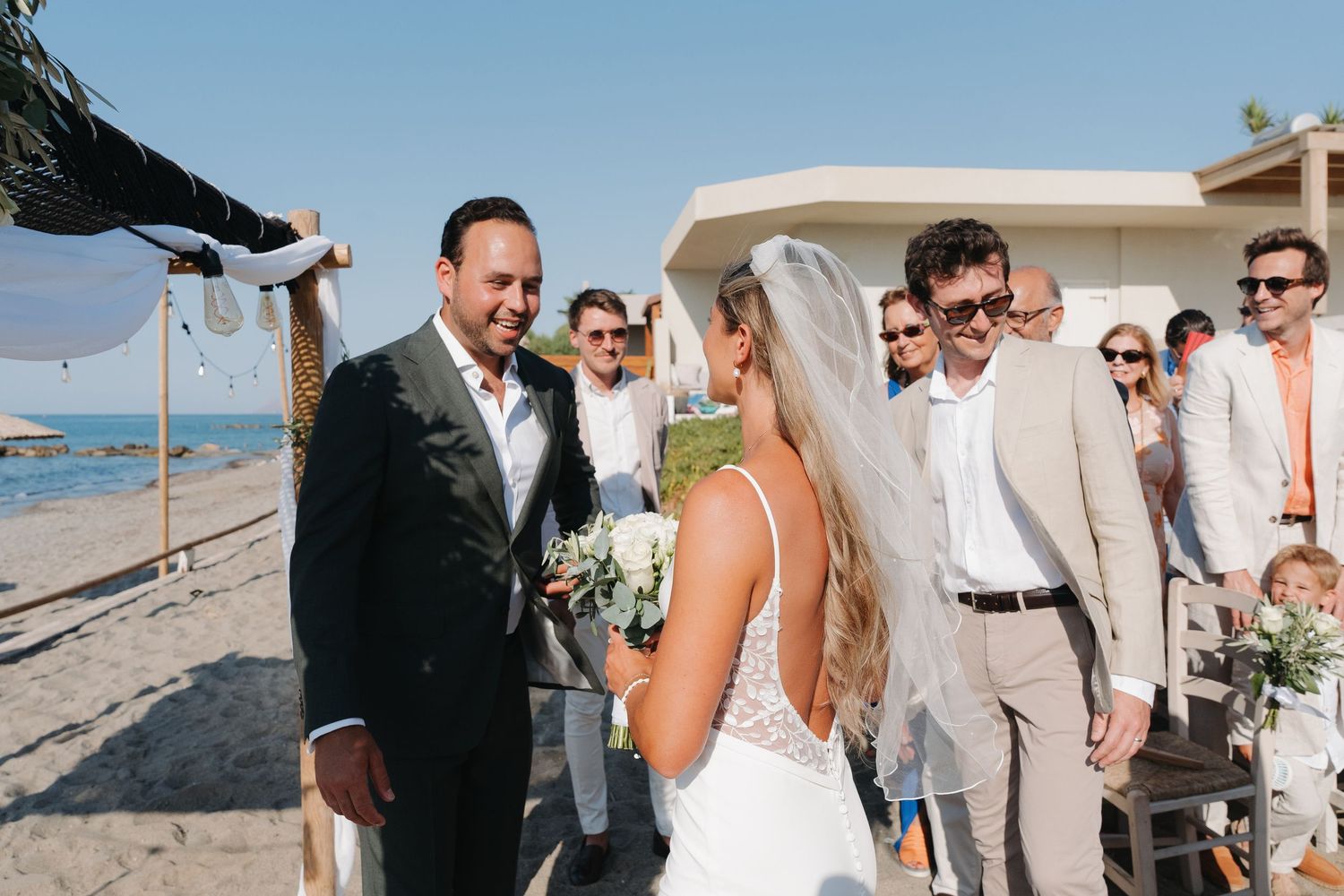 A beachfront wedding ceremony with guests gathered under sunny skies near the ocean.