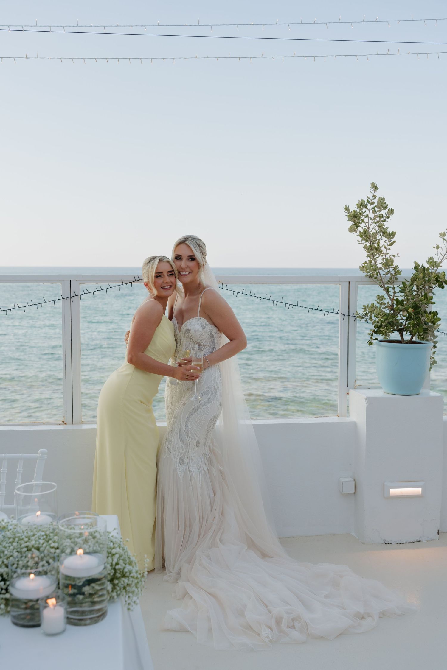Two wedding guests pose together on a white terrace overlooking the ocean.