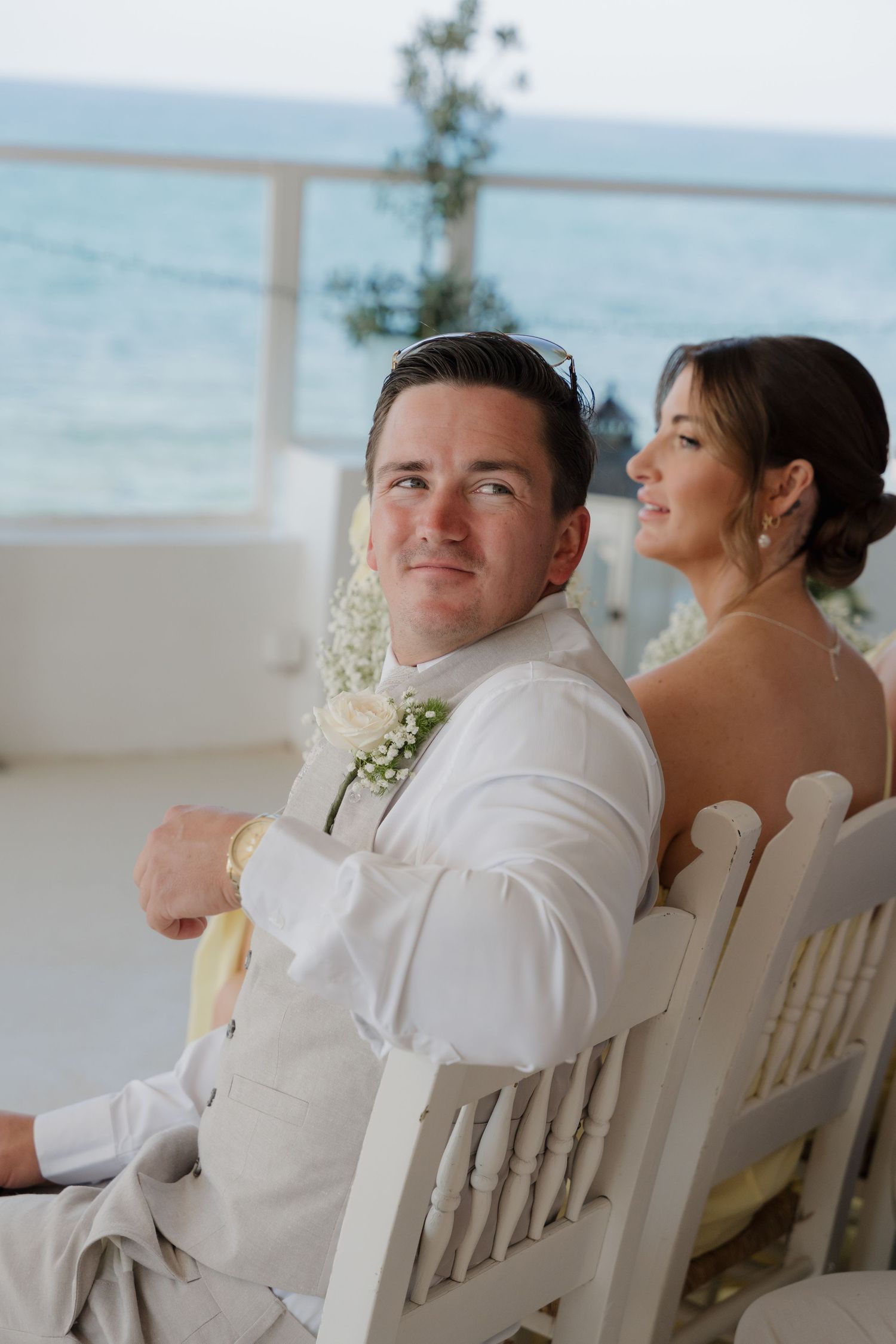 Couple seated in white chairs during beach wedding reception with ocean backdrop.