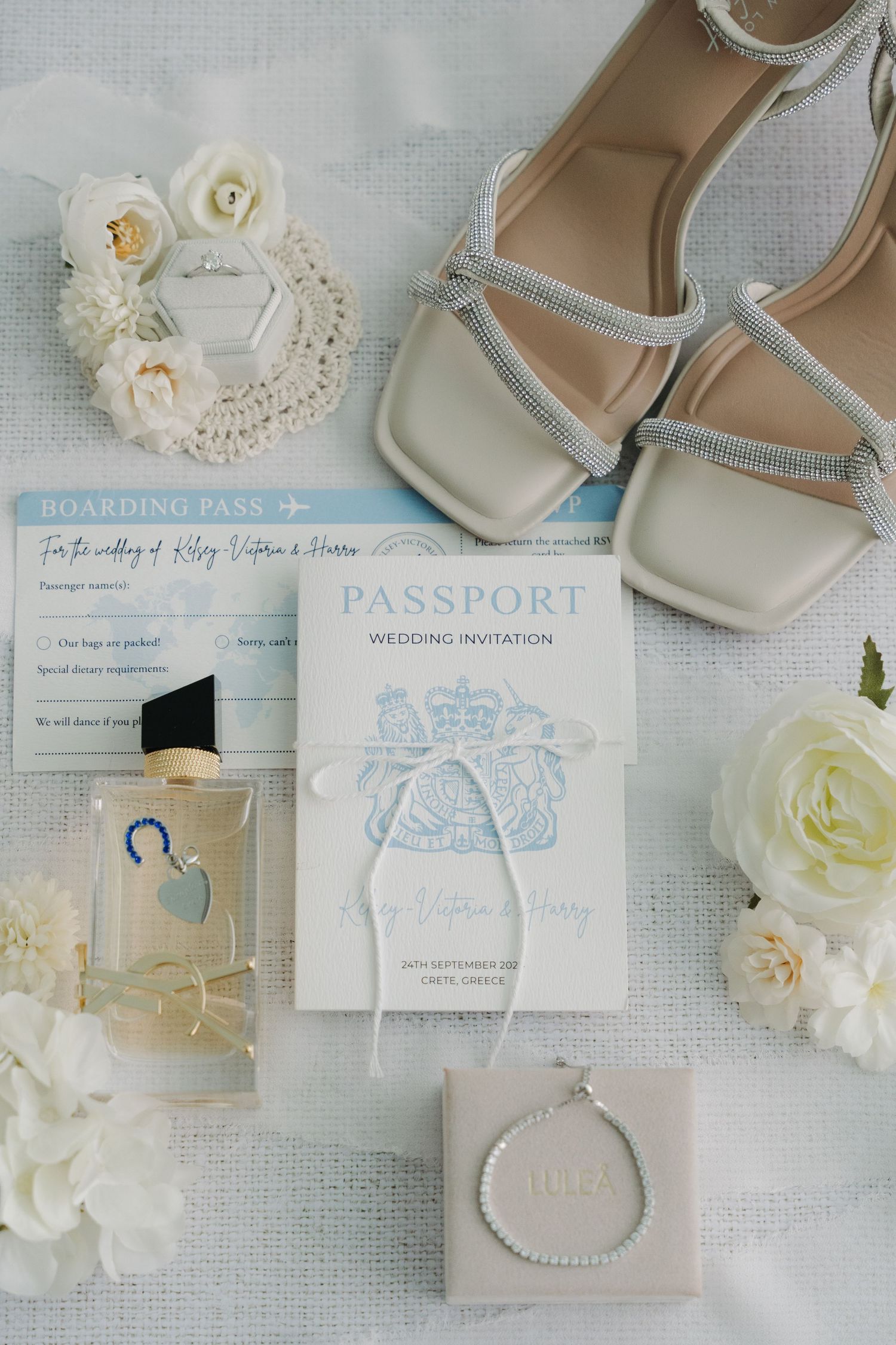 Wedding details flatlay with white shoes, passport invitation, flowers and perfume on light fabric background.