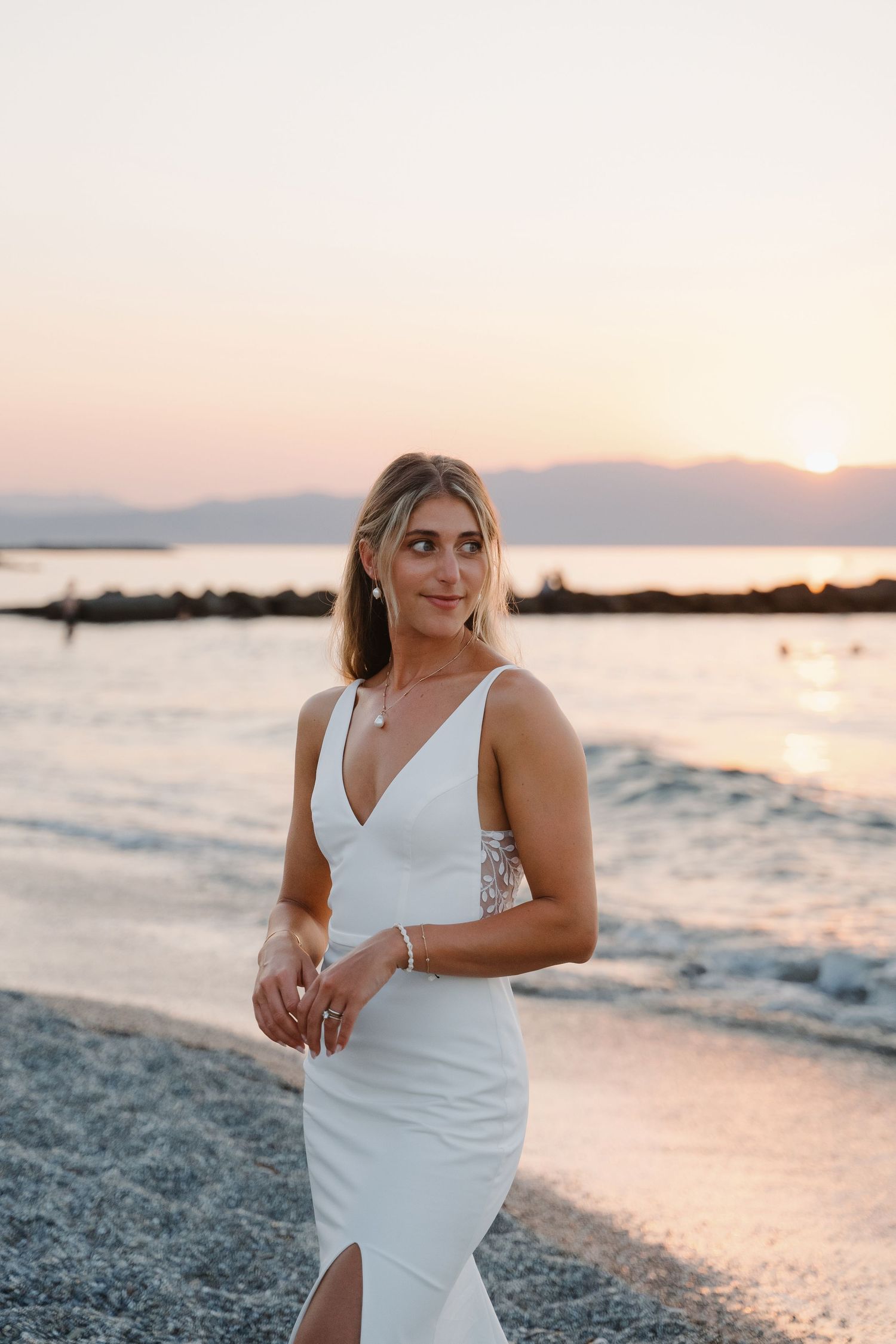 A woman in a white dress poses on a beach at sunset with mountains in the background.