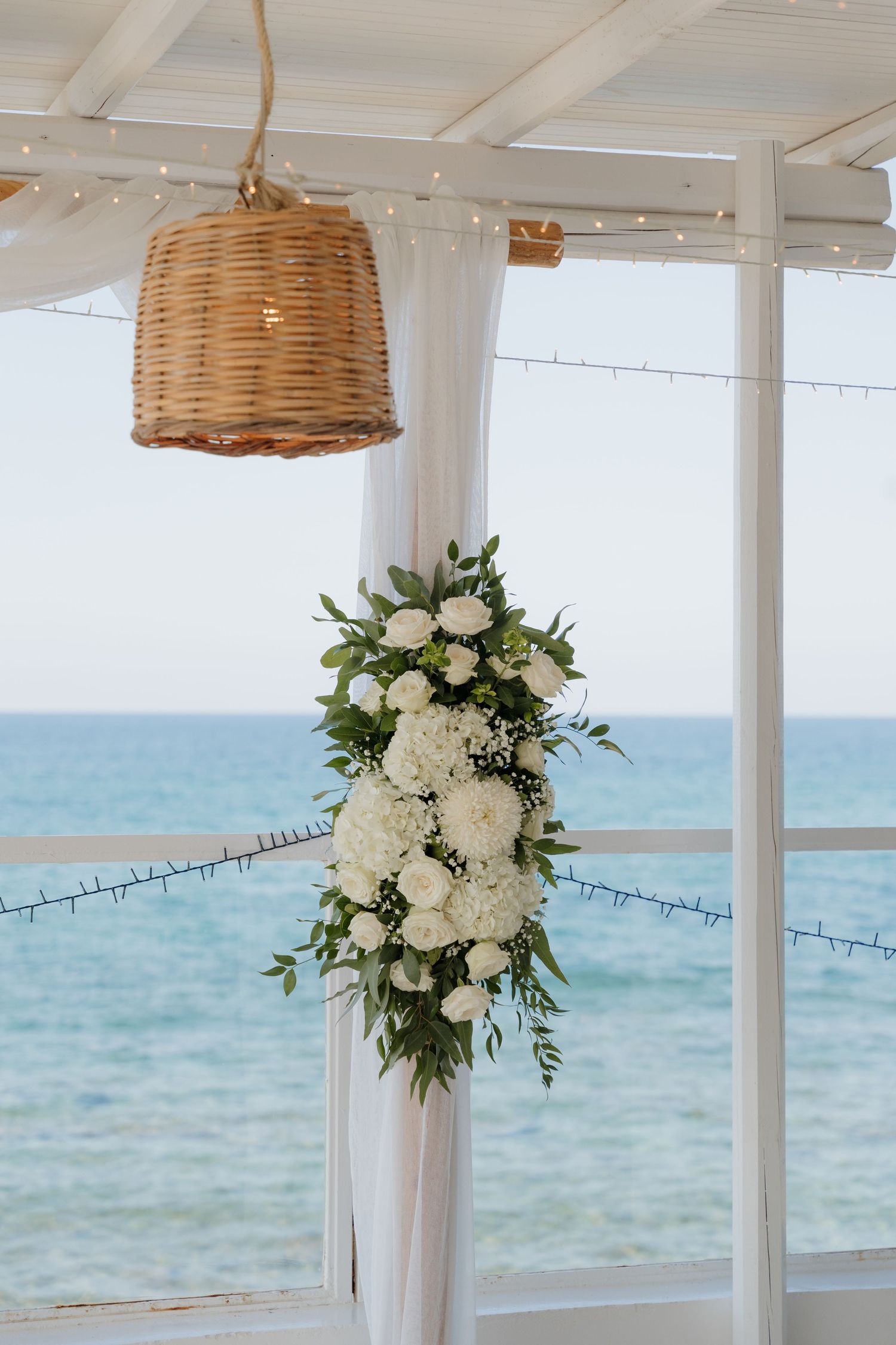 White floral arrangement with rattan pendant light hanging at a beachside wedding venue.