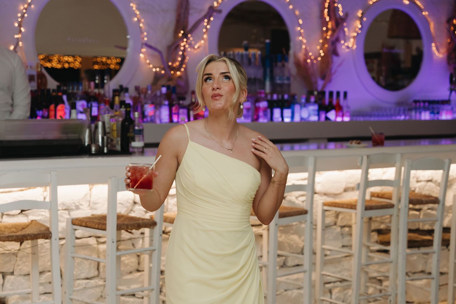 Yellow-dressed attendant poses by illuminated bar area with romantic lighting.