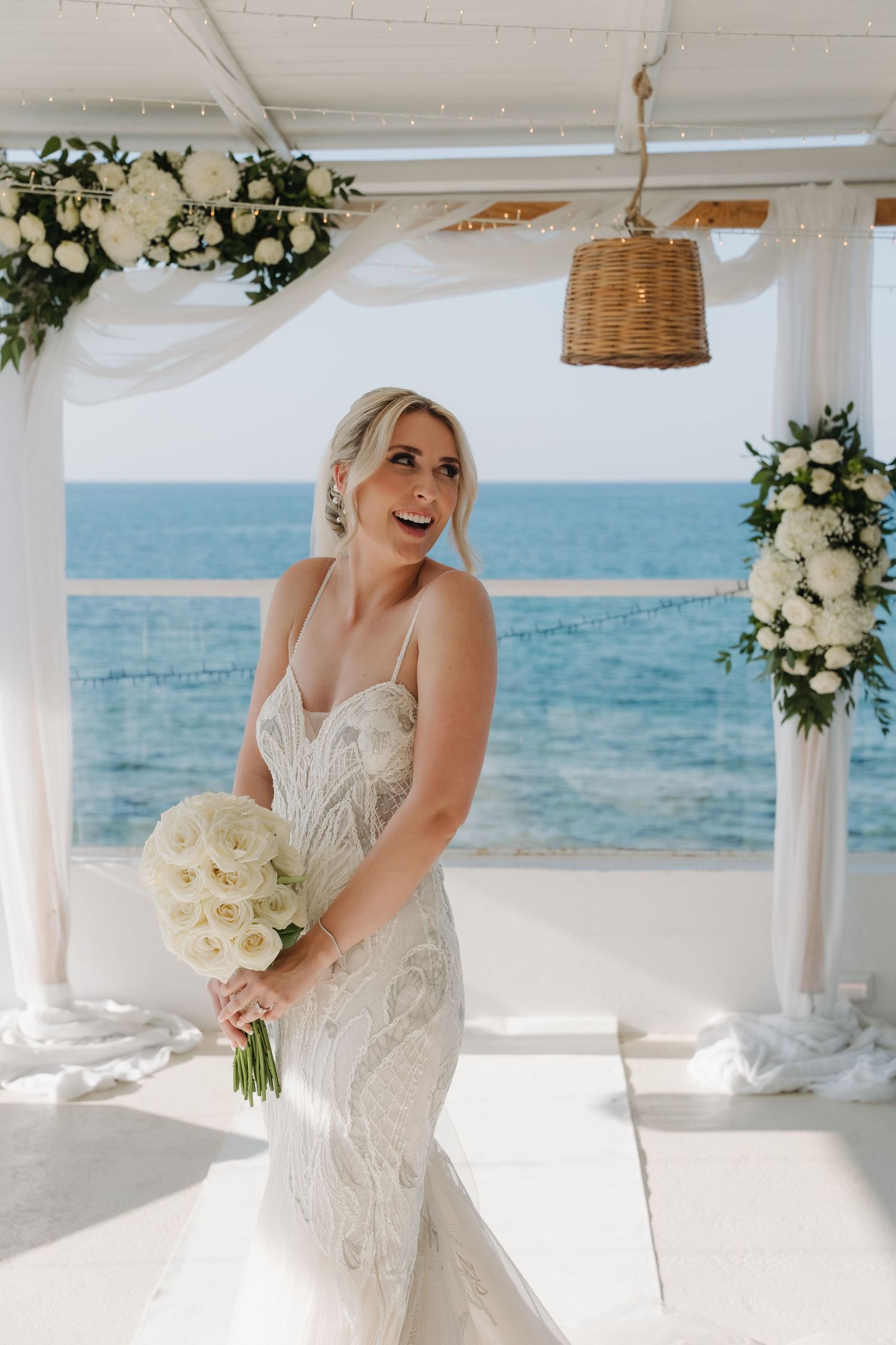 Bride in lace dress posing under decorated pergola with ocean backdrop.
