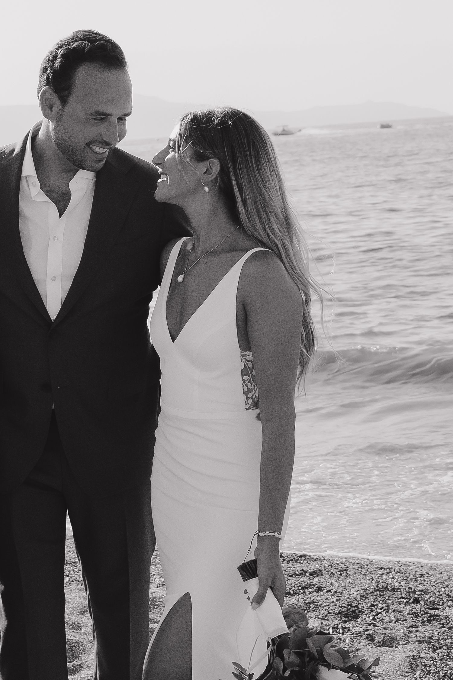 Candid seaside portrait of newlyweds sharing a laugh against ocean backdrop.