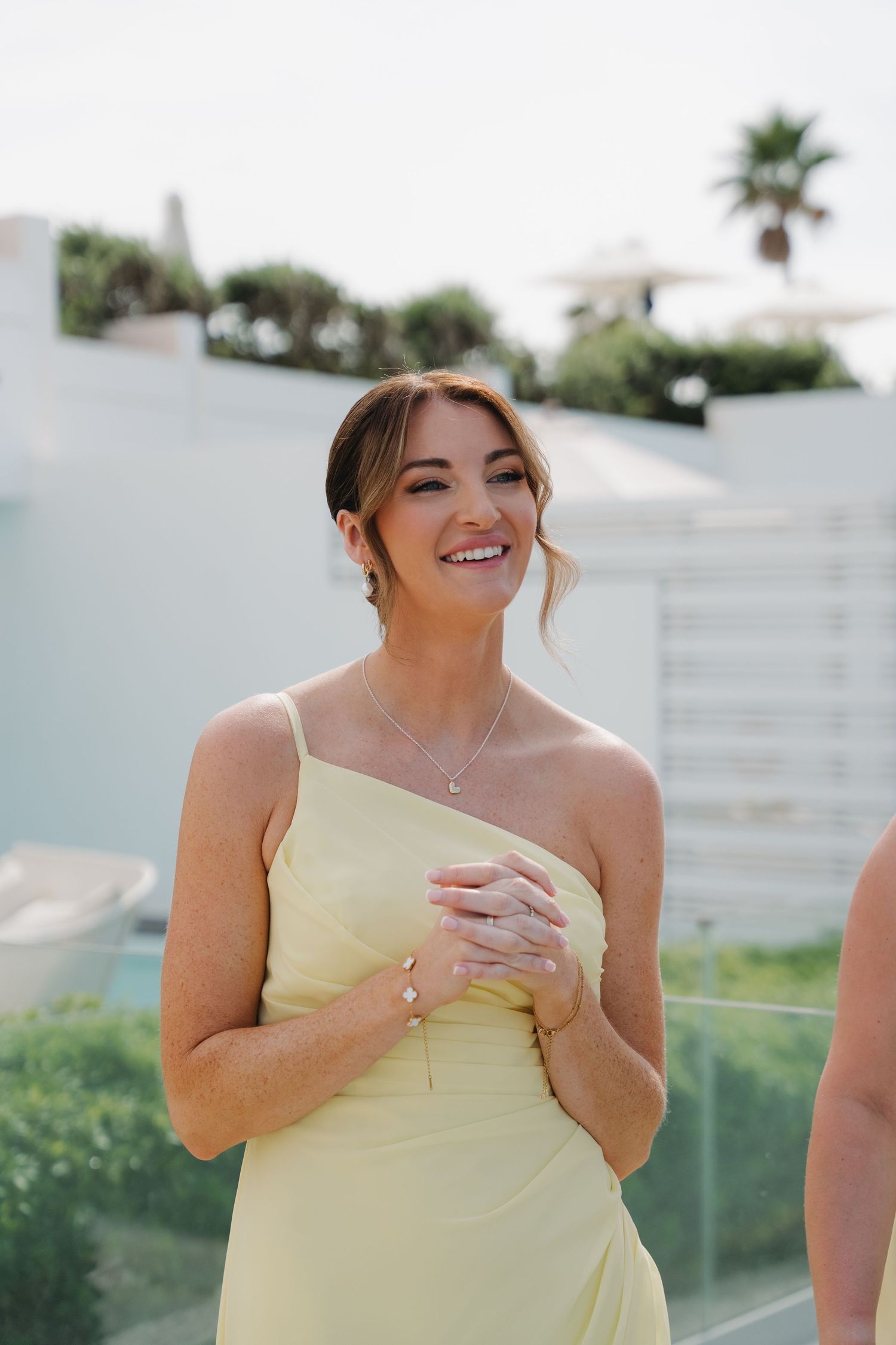 Bridesmaid in pale yellow one-shoulder dress smiling at outdoor coastal venue.