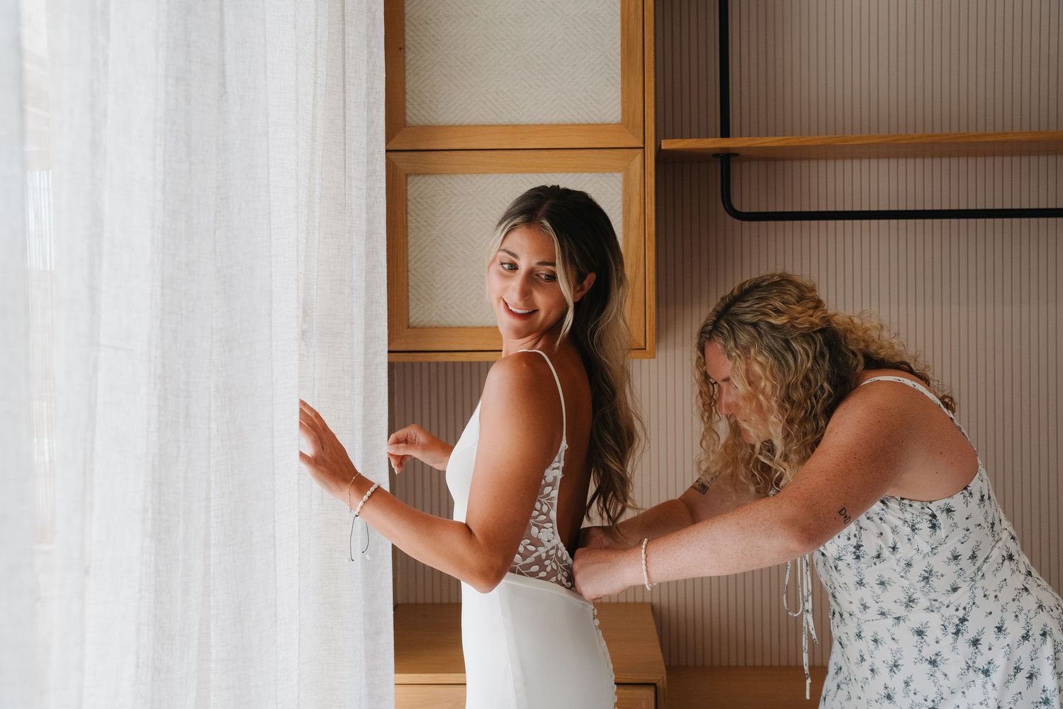 Two people in white dresses preparing for a wedding ceremony, with one helping to adjust the other's dress.