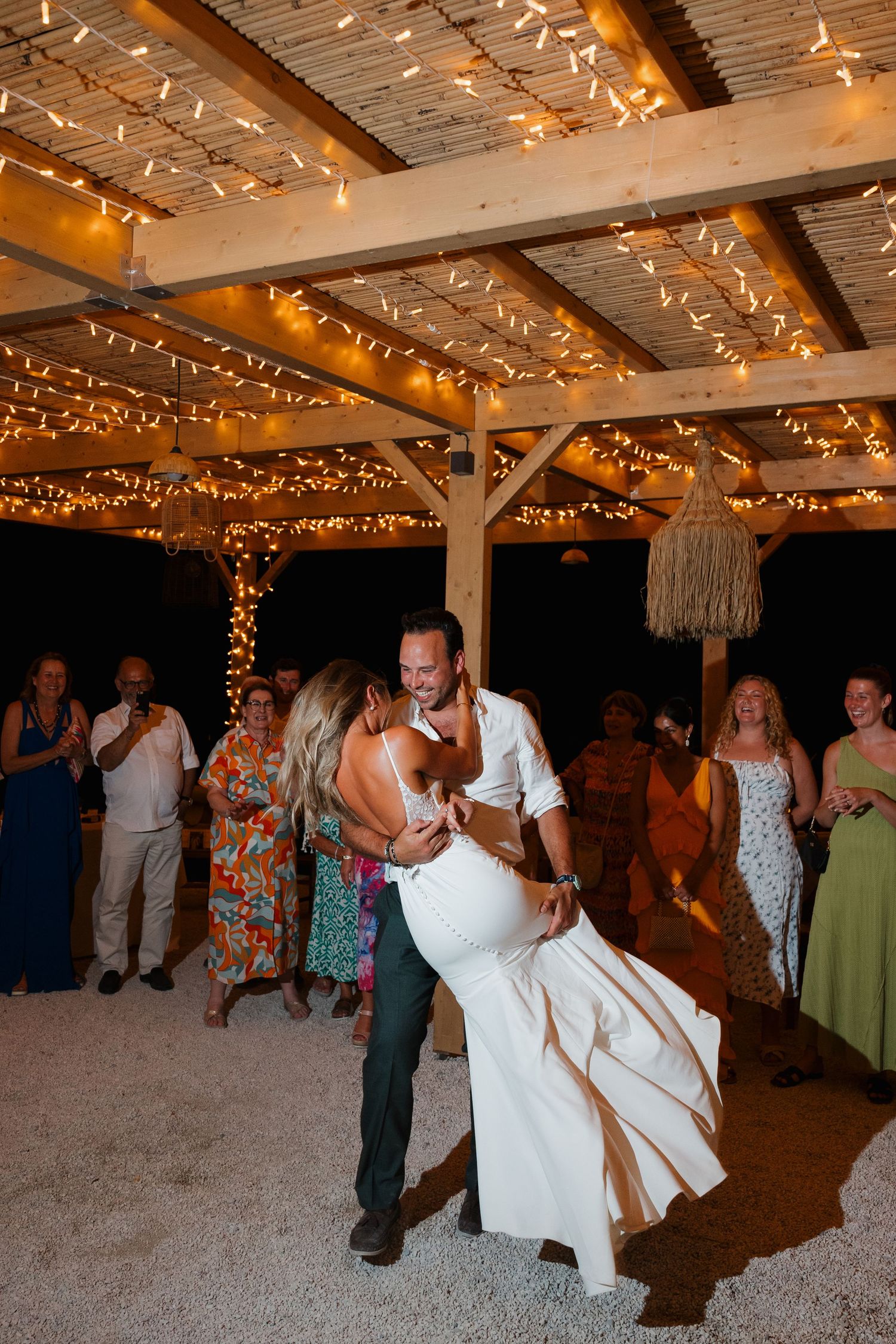 Newlyweds share their first dance under twinkling string lights at a rustic outdoor reception venue.
